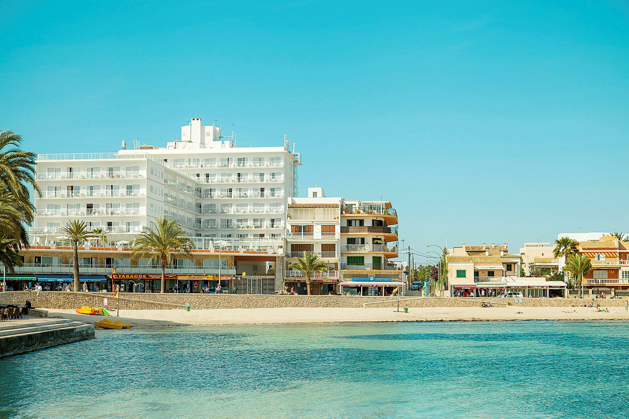 En vy över en strand med klarblått vatten i förgrunden och flera byggnader, inklusive hotell, i bakgrunden. (AI-genererad text)