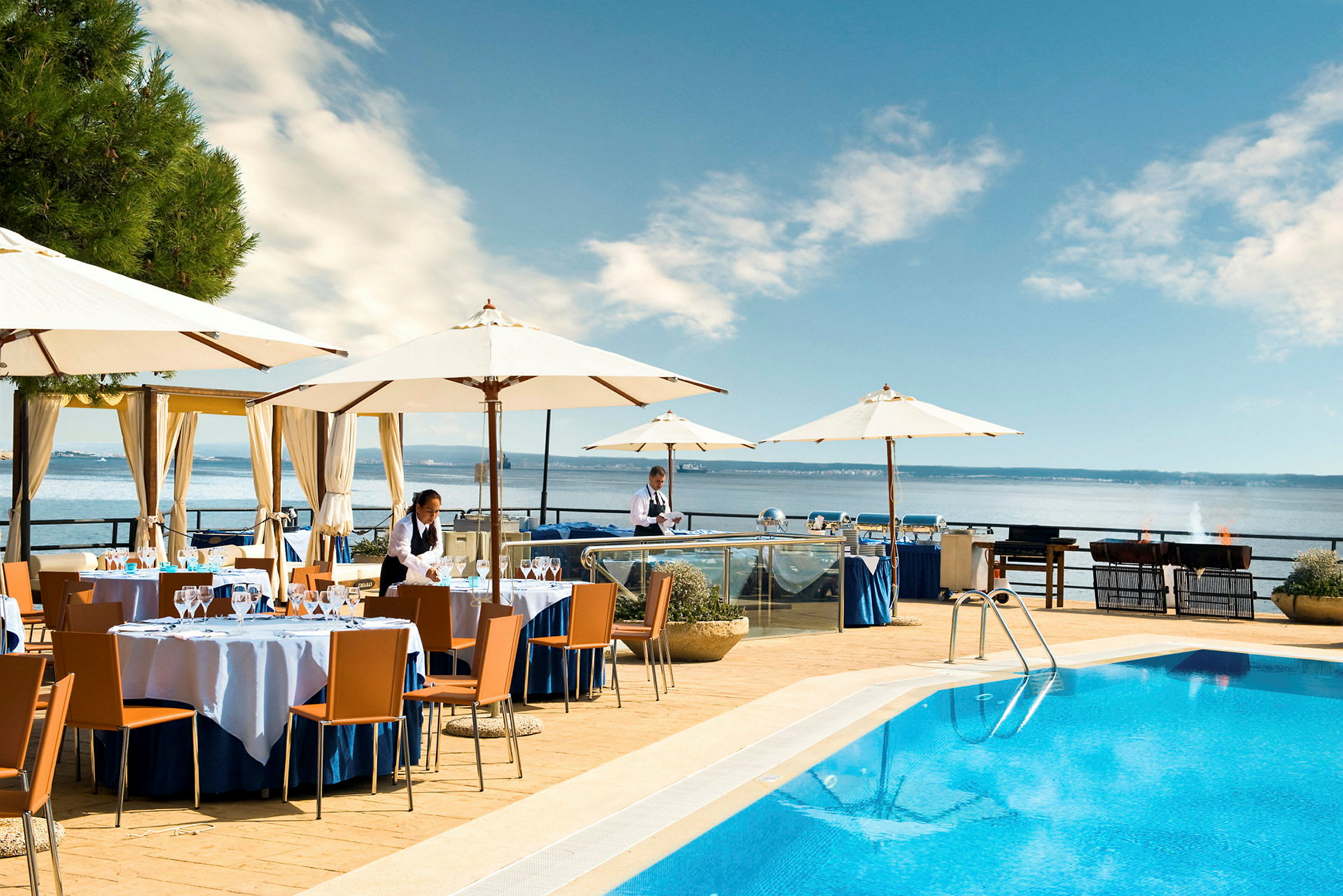Restaurangområde vid en pool med utsikt över havet. Bord och parasoller är uppställda. (AI-genererad text)