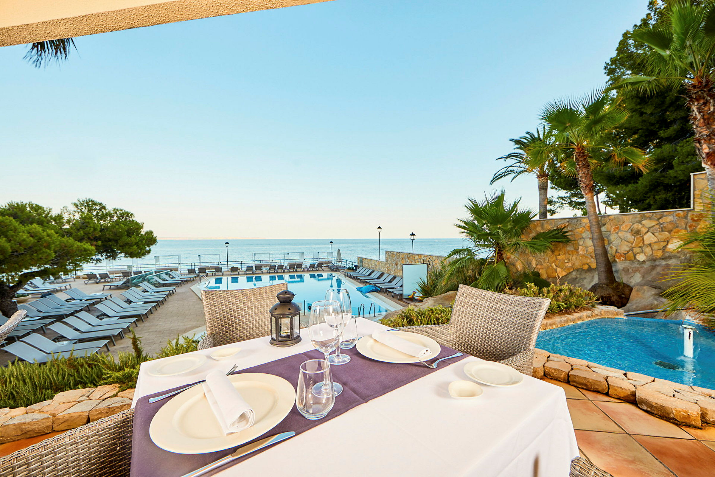 Utsikt från ett dukat bord mot en pool, solstolar och havet i bakgrunden. (AI-genererad text)