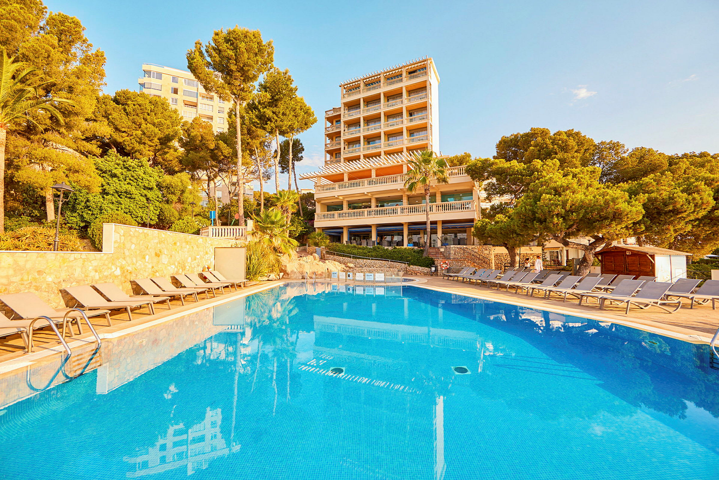 Utomhuspool framför hotellbyggnad, omgiven av solstolar och grönska. (AI-genererad text)