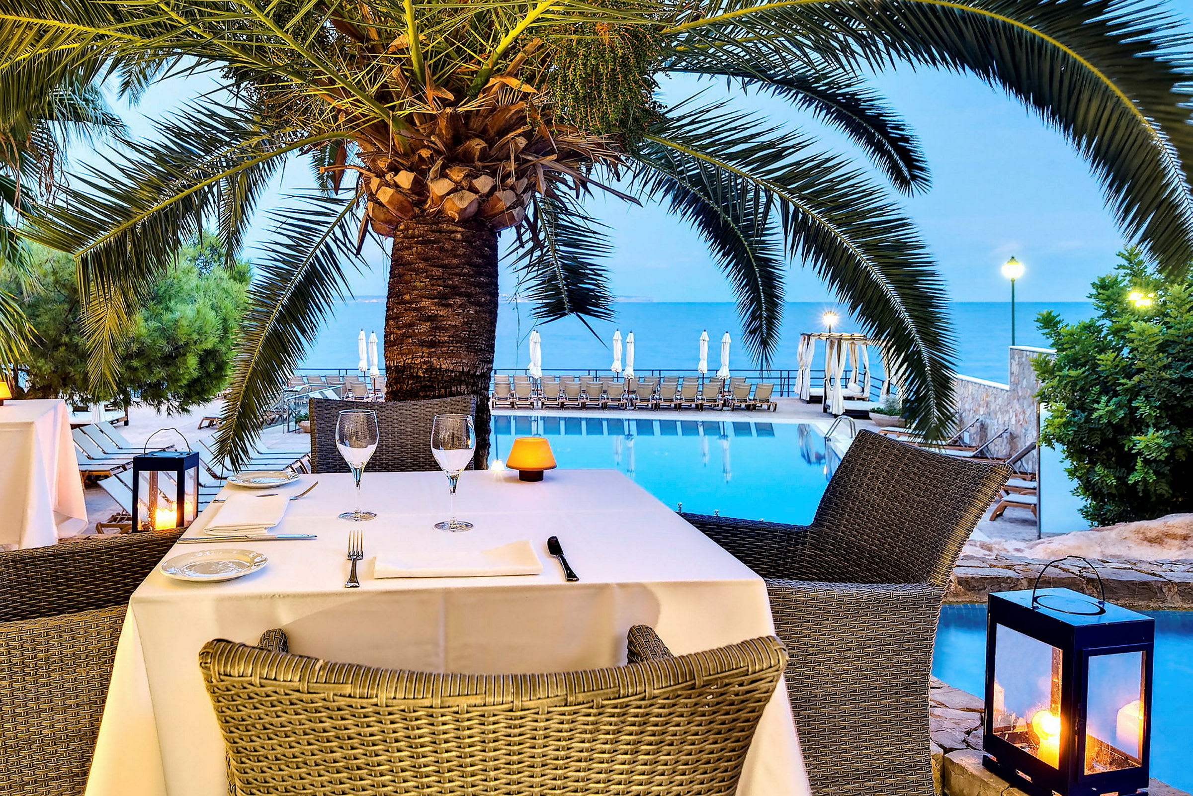 Utsikt över en restaurang vid en pool nära havet med en palm i förgrunden. (AI-genererad text)