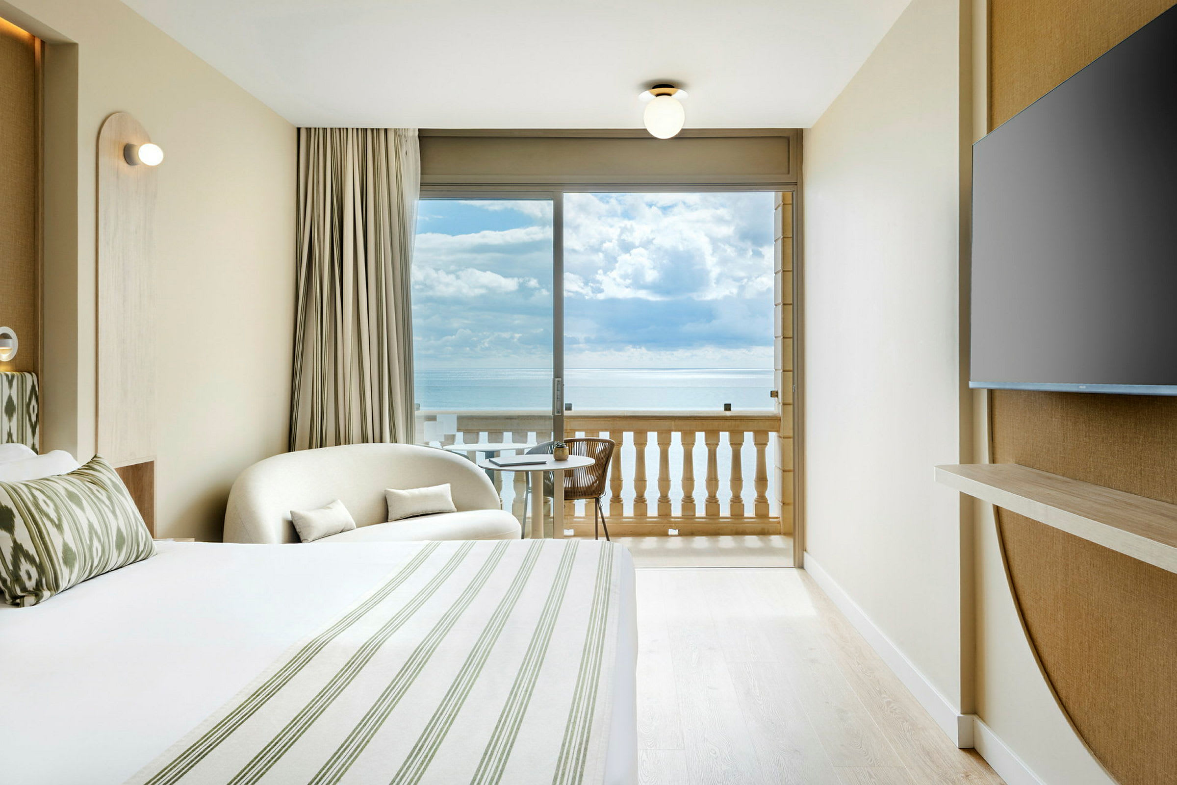 Ett ljust hotellrum med en säng, en sittplats, ett TV på väggen och en balkong med utsikt över havet. (AI-genererad text)
