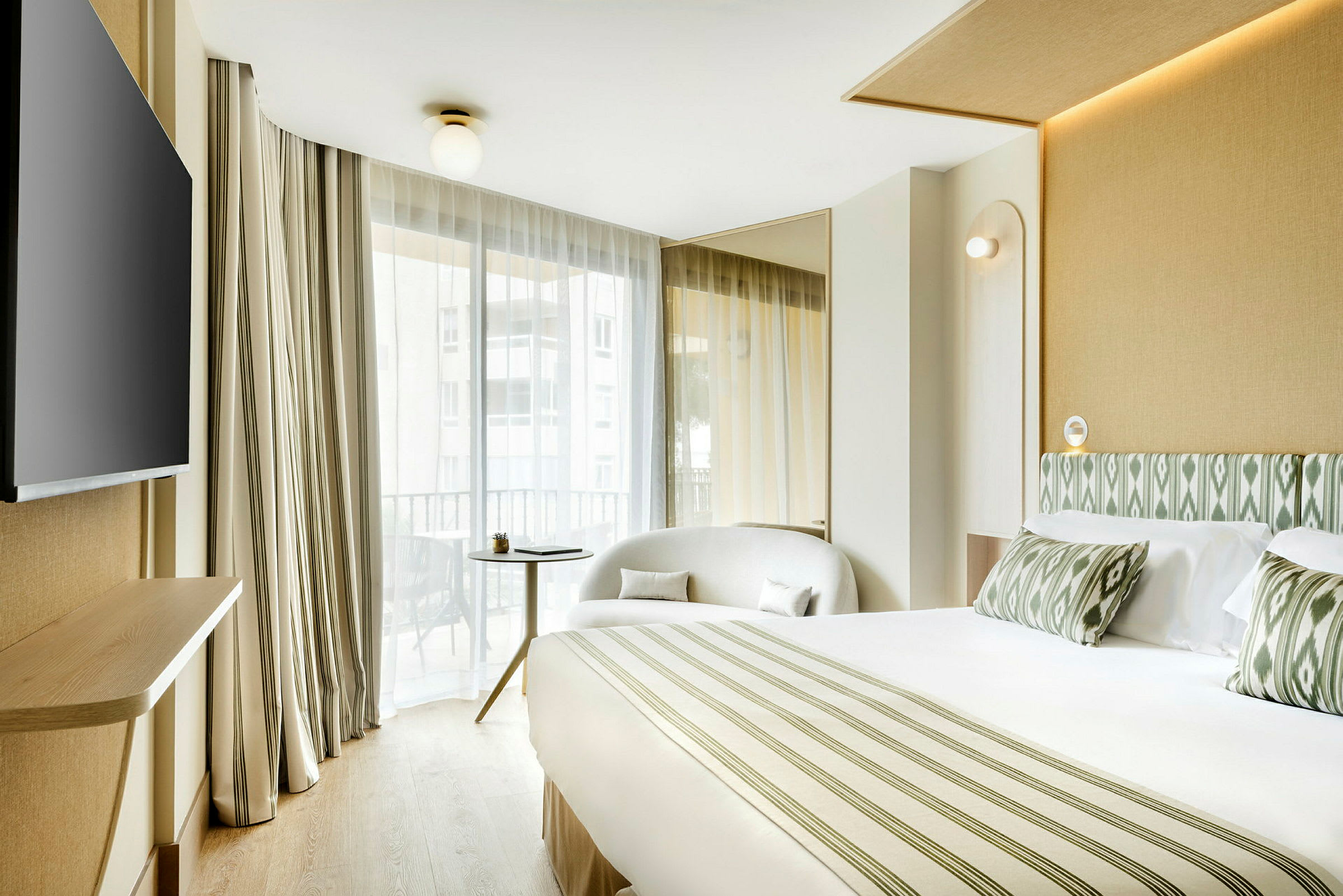 Ett hotellrum med en stor säng, fåtölj, bord och en TV, samt stora fönster med ljusa gardiner. (AI-genererad text)