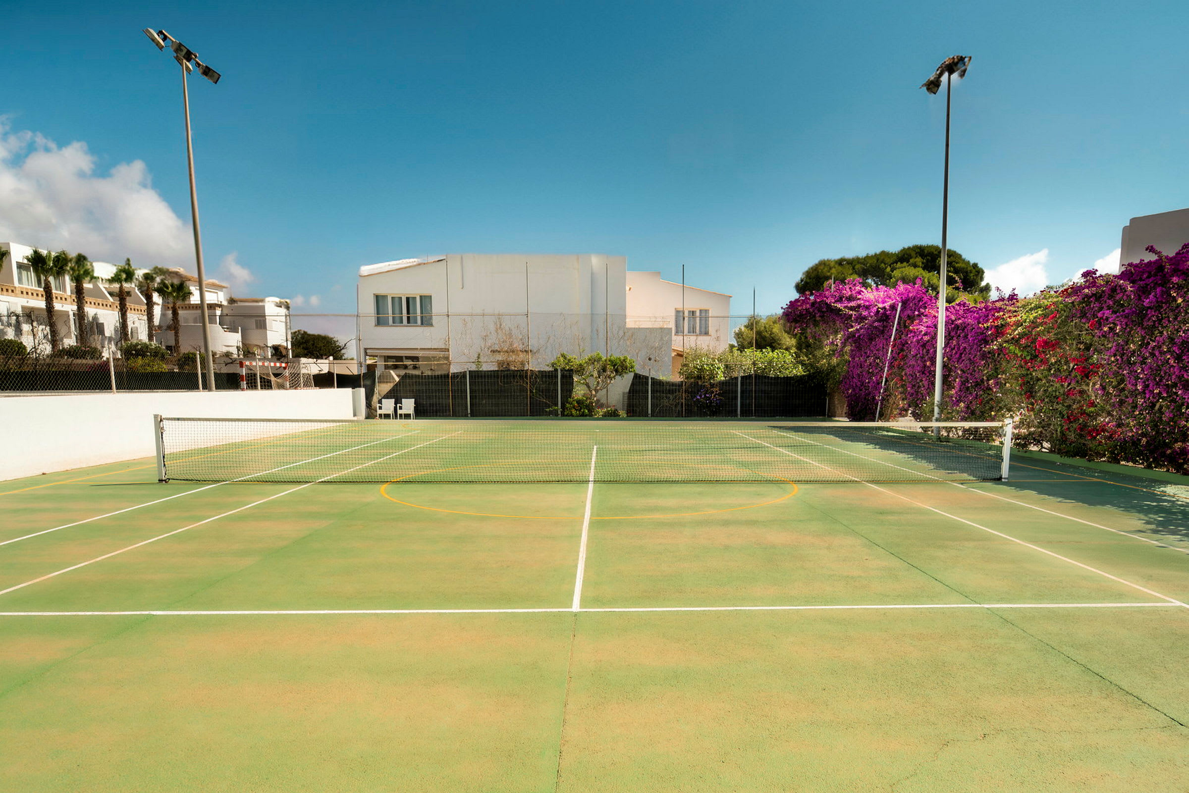 En tennisplan med grönt underlag omgiven av byggnader och växtlighet. (AI-genererad text)