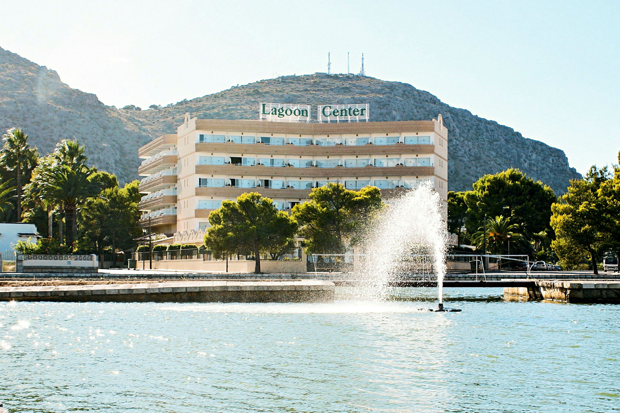 Hotell Lagoon Center vid strandpromenaden med en fontän och berg i bakgrunden. (AI-genererad text)