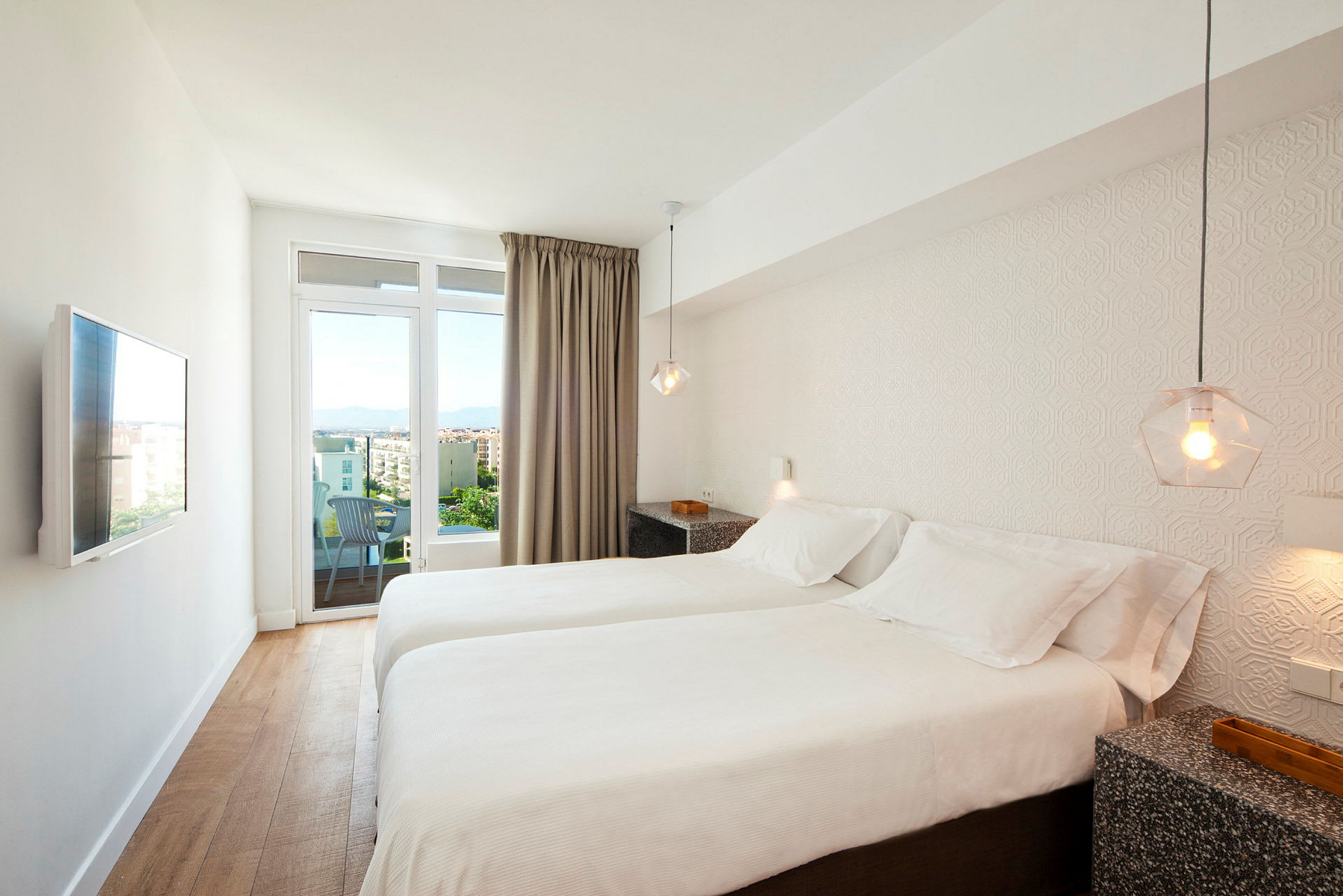 Ett hotellrum med två enkelsängar, vita sängkläder, en vägg-TV och utsikt över en stad genom glasdörrar. (AI-genererad text)