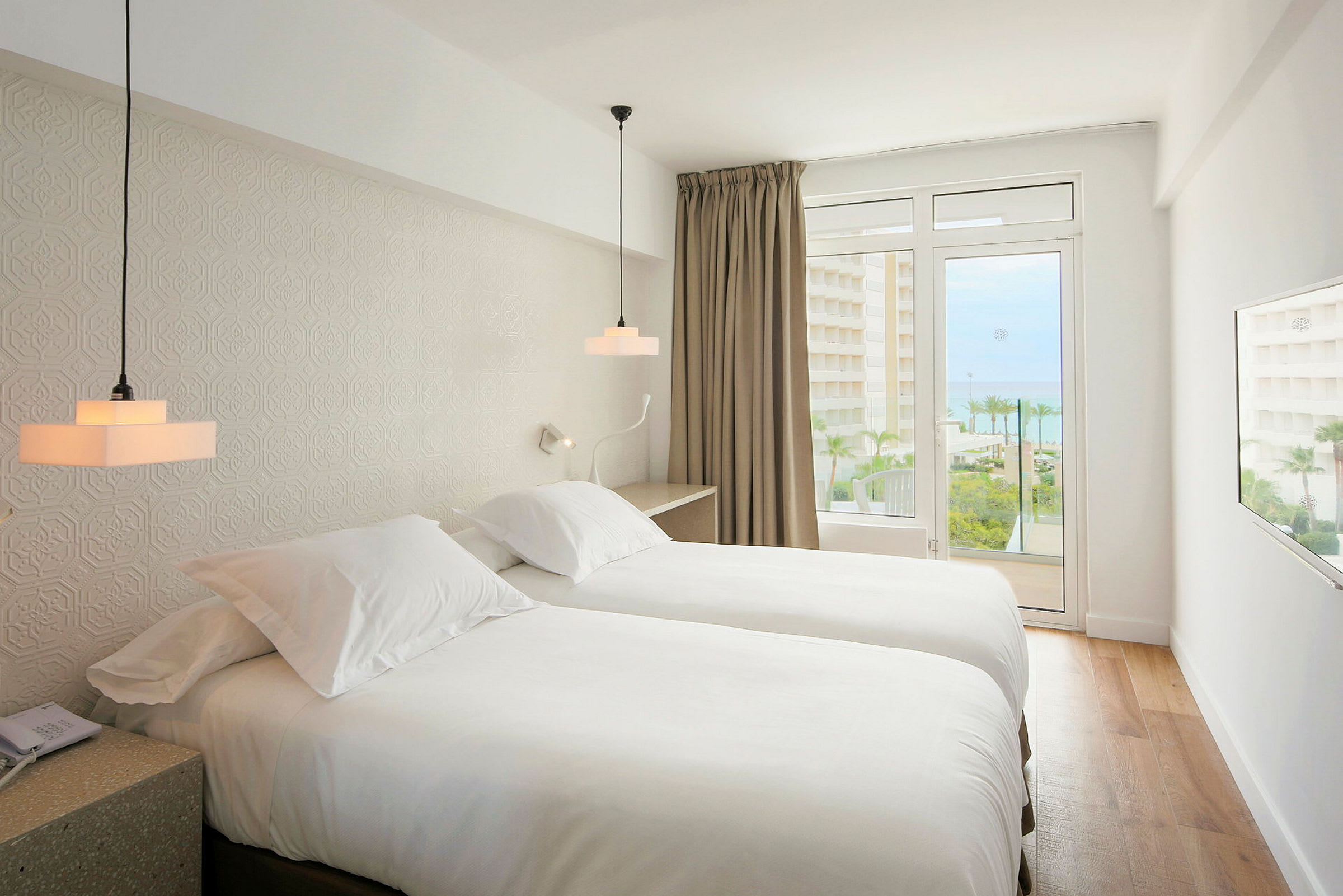 Ett hotellrum med två enkelsängar, ljus inredning och en balkong med utsikt mot havet. (AI-genererad text)
