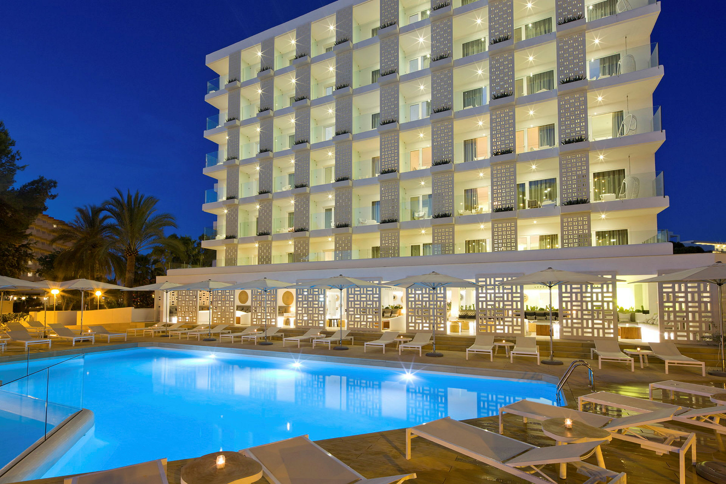 Bild av ett hotell med en upplyst pool, solstolar och en modern fasad kvällstid. (AI-genererad text)