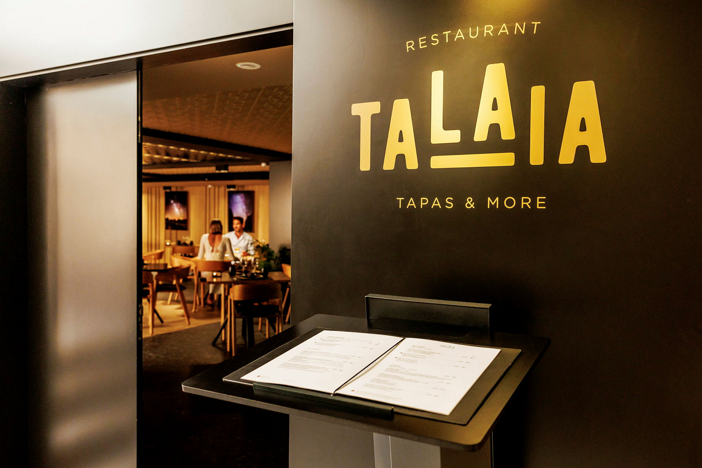 Restaurang Talaia med en meny på ett stativ nära ingången, som visar en inblick i en stilfull interiör med sittplatser. (AI-genererad text)