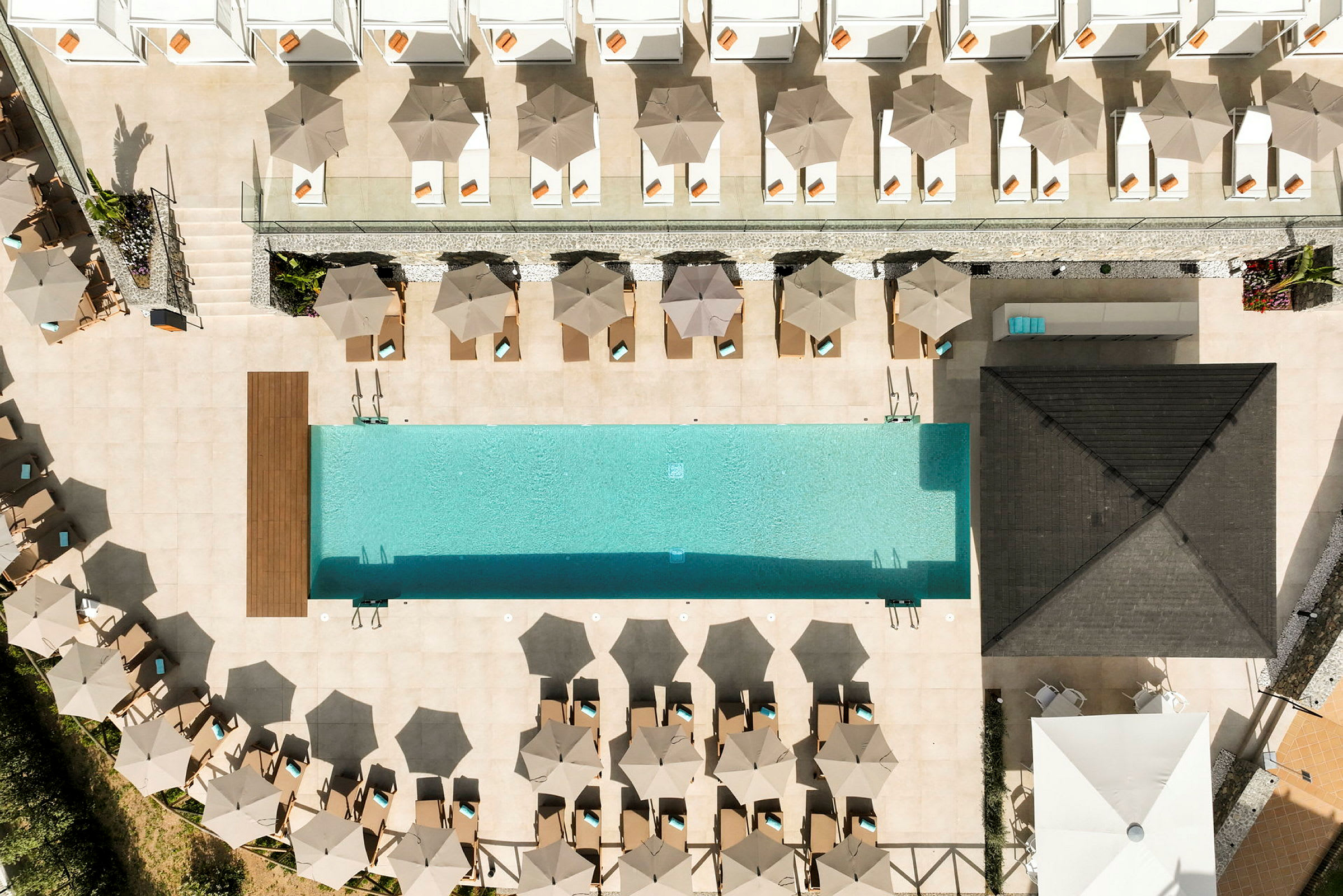 Flygfoto av en rektangulär pool med solstolar och parasoller runt omkring. (AI-genererad text)