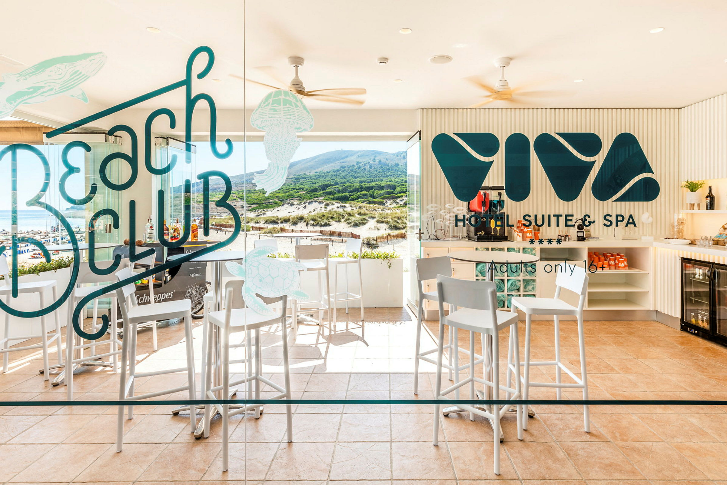 Bild på en hotellbar med höga stolar, bord och glasvägg med texten 'Beach Club' och 'Viva Hotel Suite & Spa' samt utsikt mot en strand och grönskande höjder. (AI-genererad text)