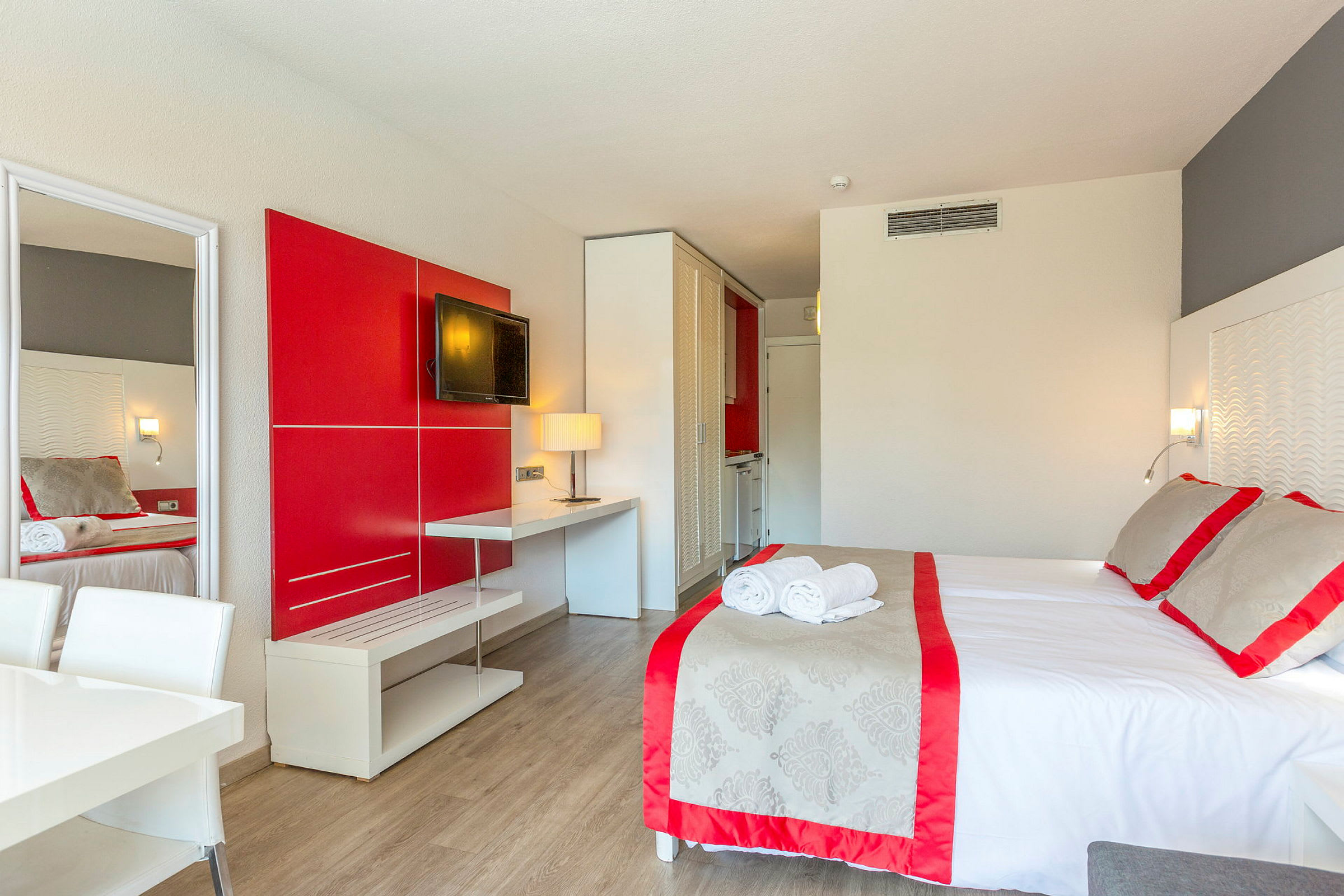 Ett hotellrum med en säng, röda och grå detaljer, en TV på väggen och ett bord med en lampa. (AI-genererad text)