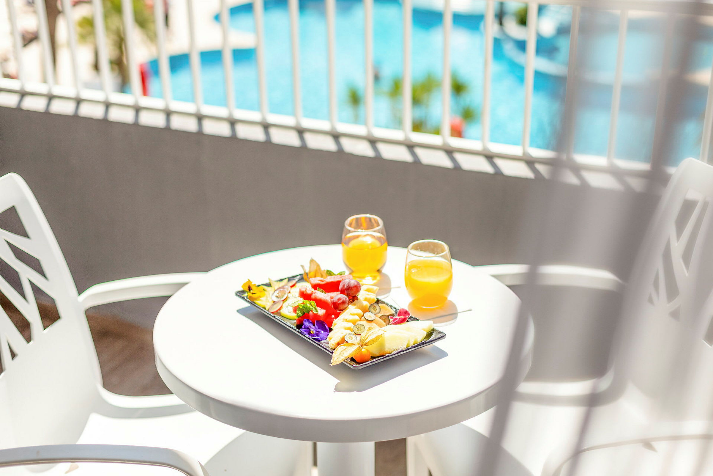 En vit bord med färsk frukt och två glas apelsinjuice, med en pool i bakgrunden. (AI-genererad text)