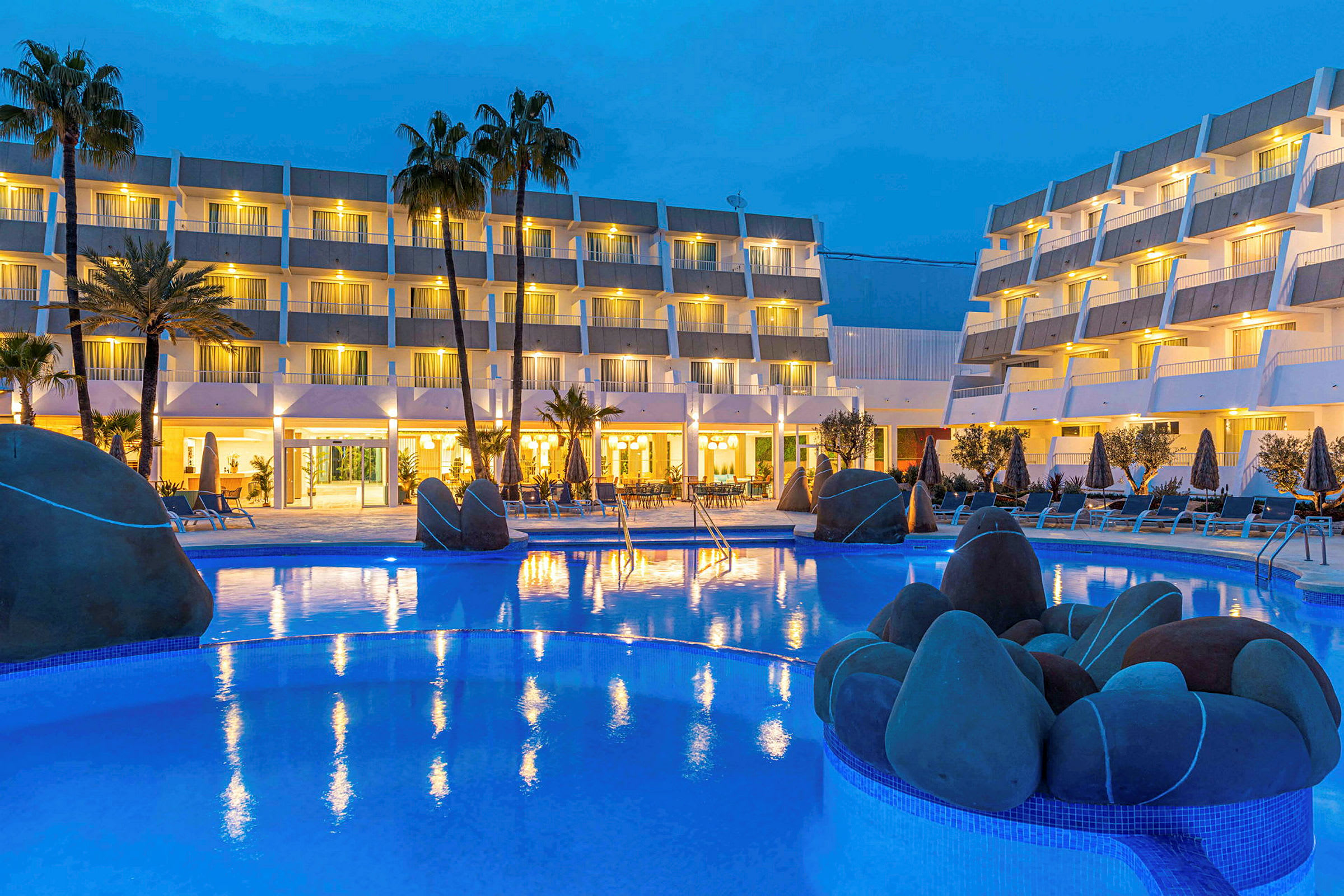 Hotellfasad i skymningen med upplyst poolområde och palmer. (AI-genererad text)