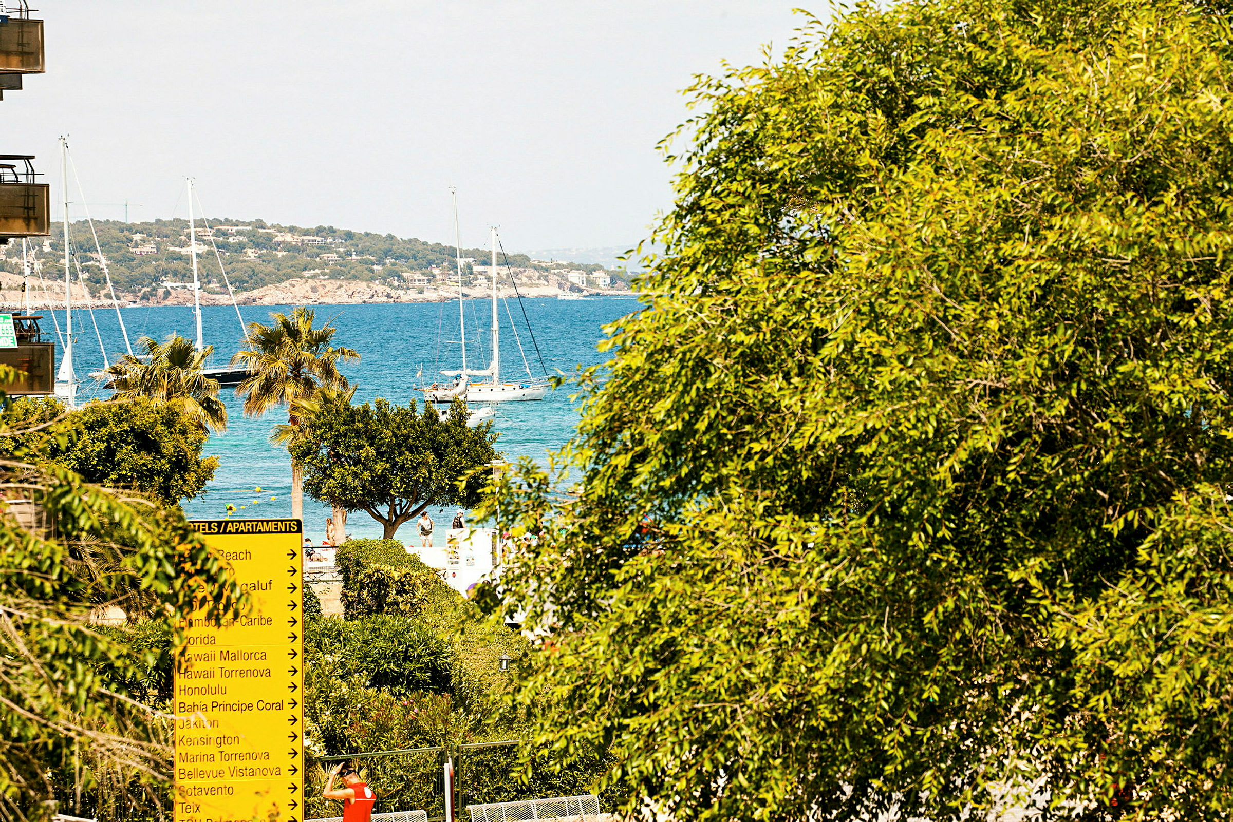 Vy över ett kustlandskap med båtar på havet, omgiven av träd och en skylt som pekar mot olika hotell och lägenheter. (AI-genererad text)