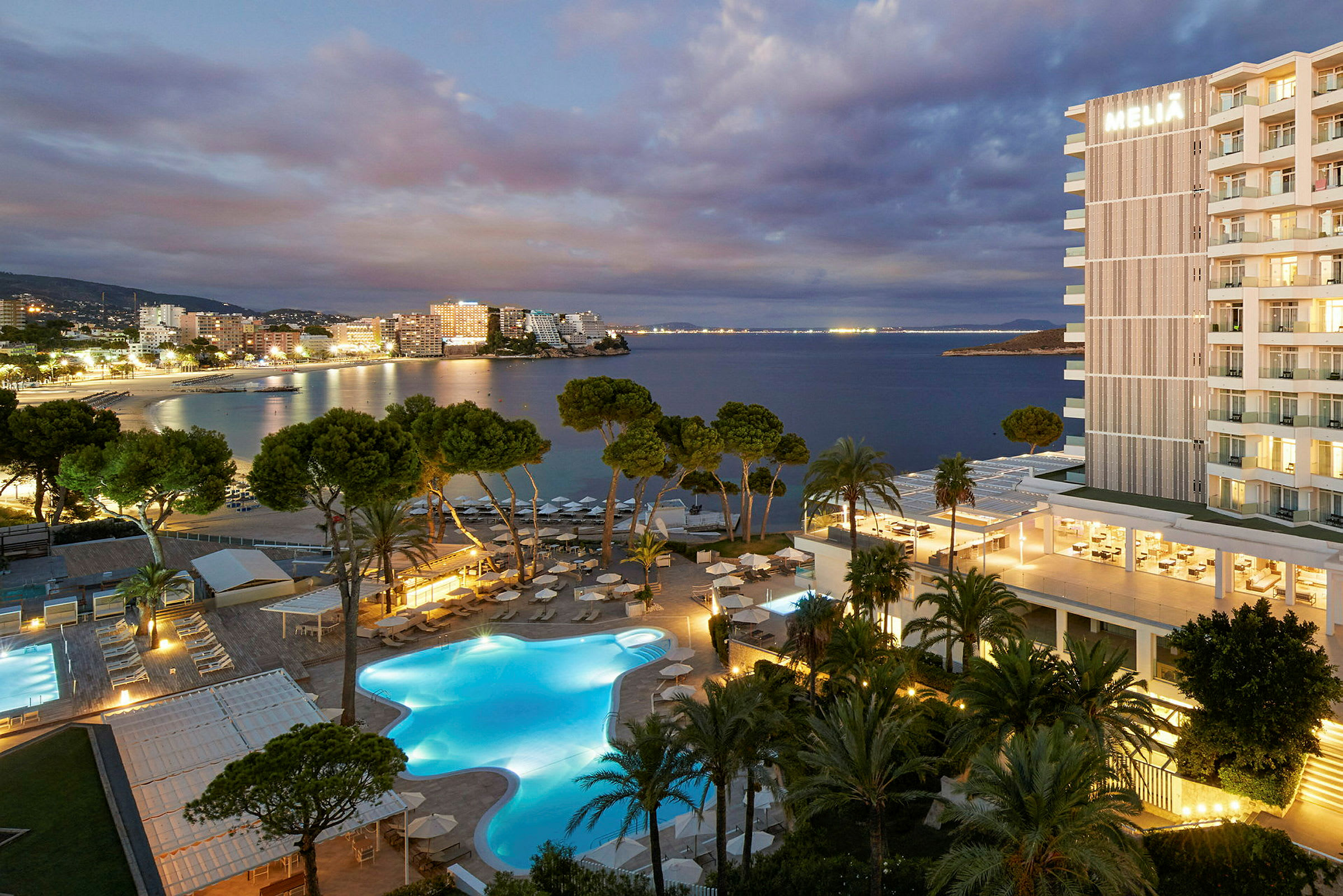 Kvällsutsikt över en belyst hotellbyggnad med poolområde i förgrunden och havet i bakgrunden. (AI-genererad text)