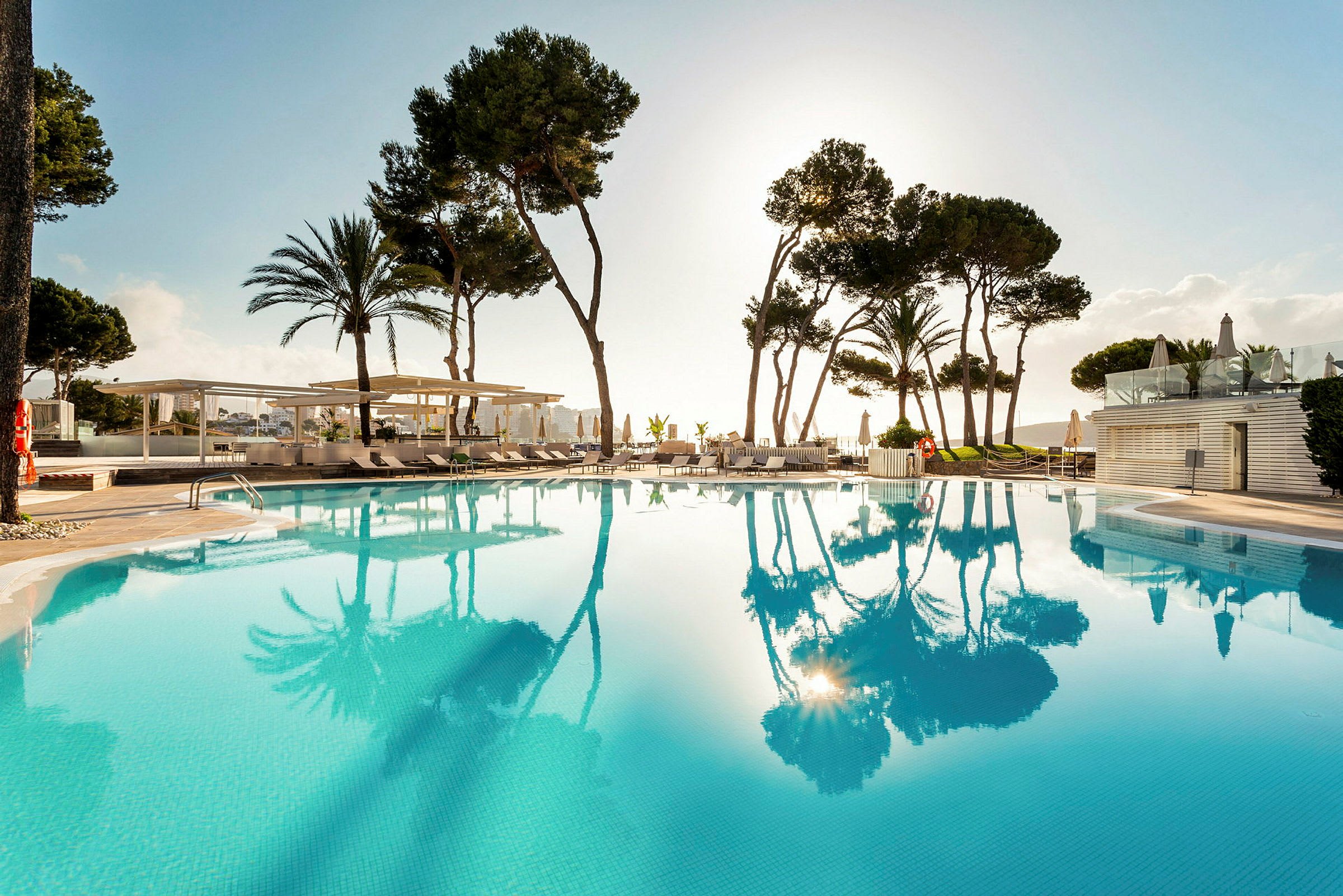Bild av en pool med klart vatten, omgiven av höga träd och solstolar. Solen syns i horisonten. (AI-genererad text)