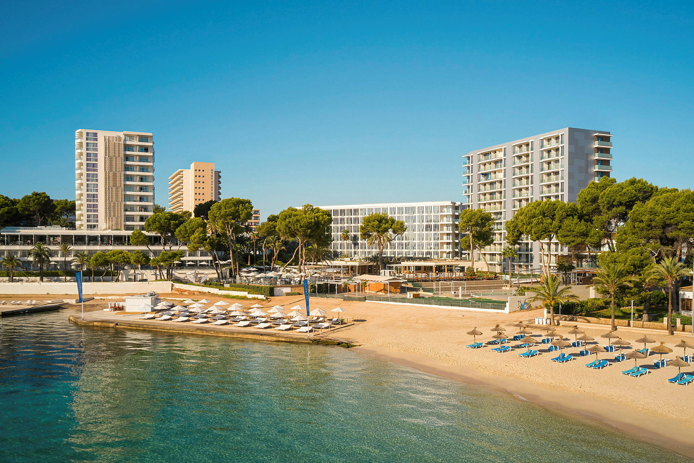 En strand med solsängar och parasoll bredvid hotellbyggnader och grönska. (AI-genererad text)