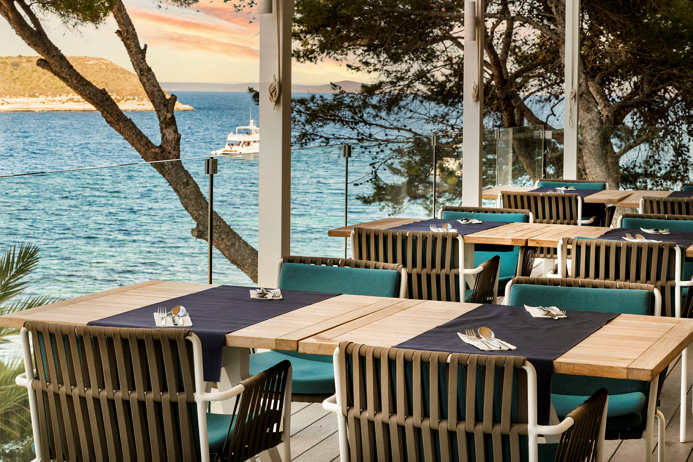 Uteservering med dukade bord med utsikt över havet och träd. (AI-genererad text)