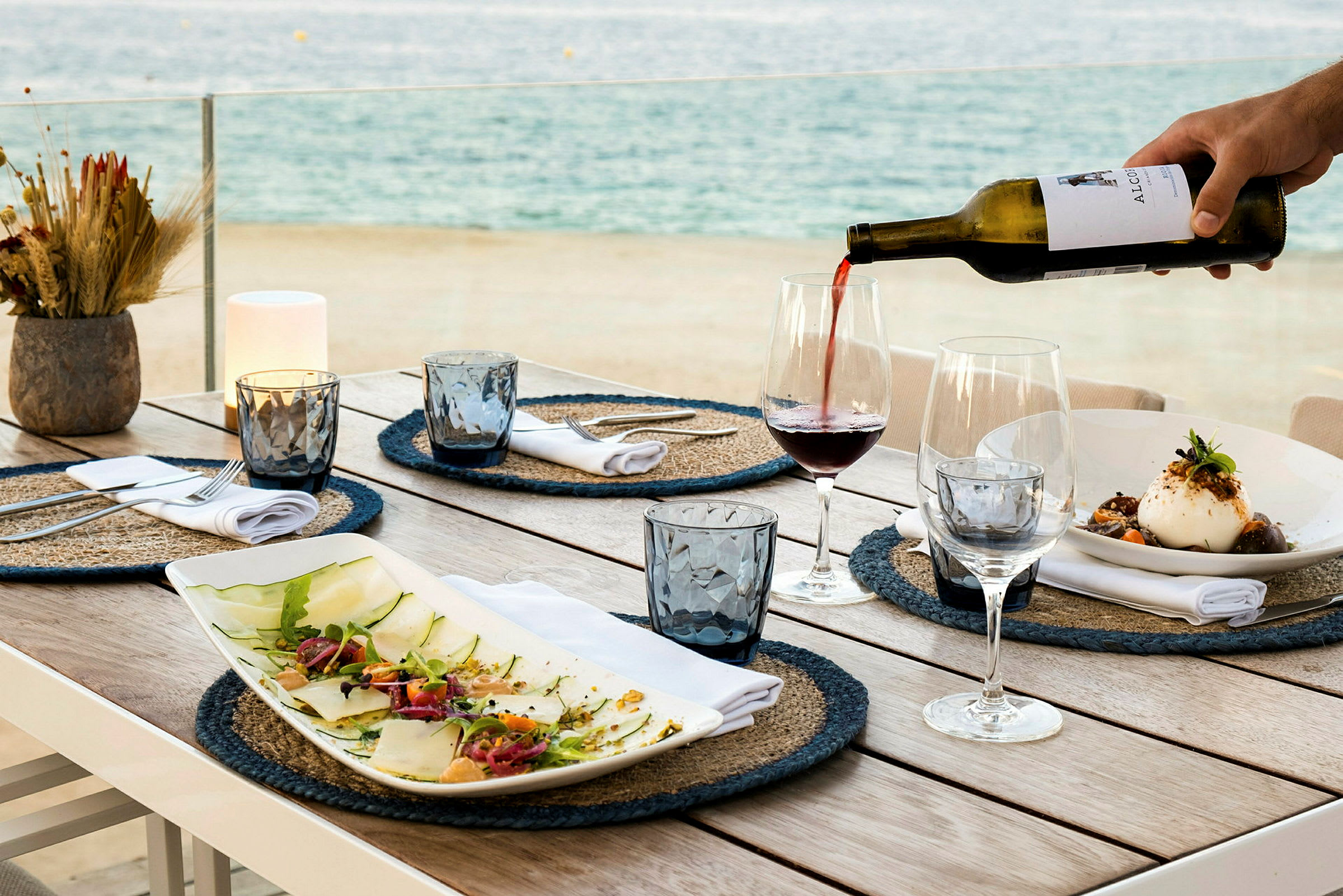 Ett dukat bord med tallrikar, glas och flaskor. Vin hälls upp i ett glas med havet i bakgrunden. (AI-genererad text)