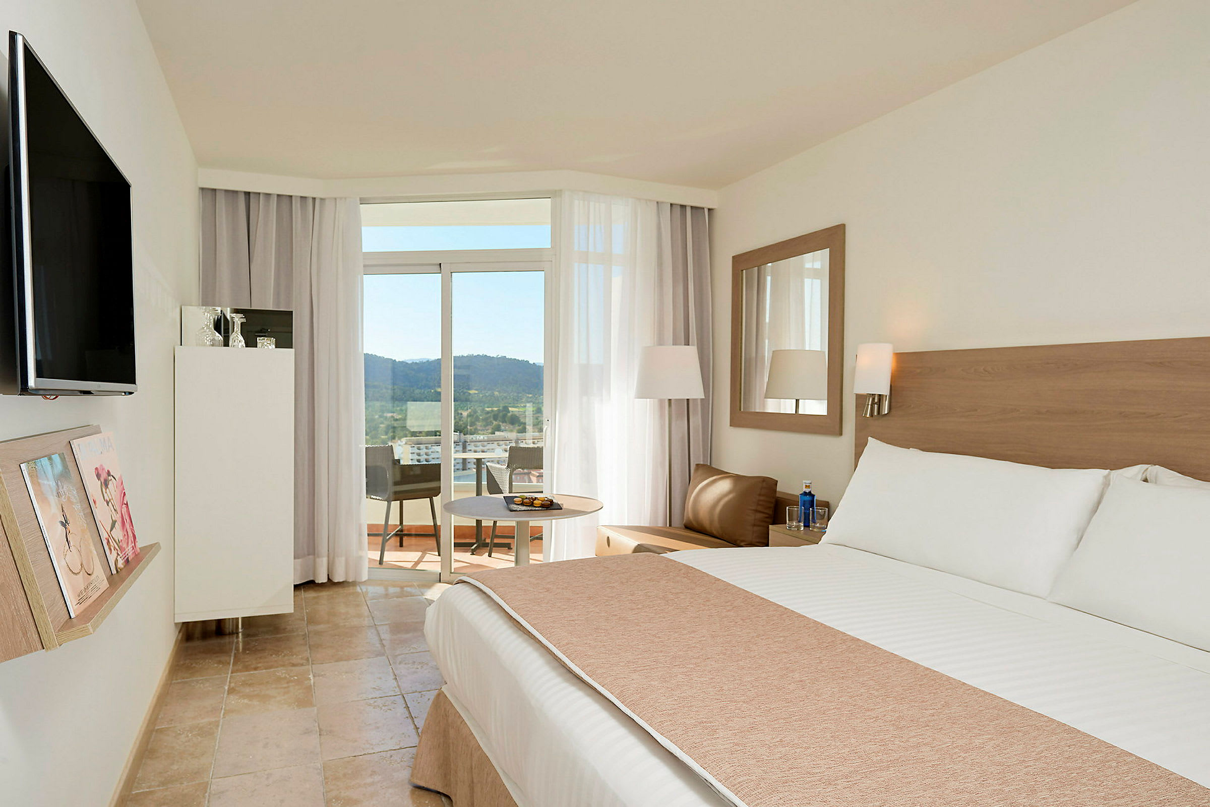 Ett hotellrum med en stor säng, sittgrupp och utgång till en balkong med utsikt över träd och landskap. (AI-genererad text)