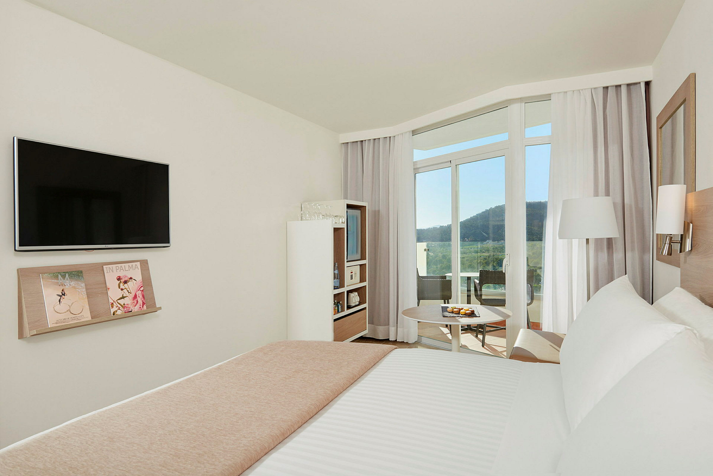 Ett ljust och modernt hotellrum med en dubbelsäng, TV på väggen och en hylla med tidningar. Rummet har stora fönster och en balkong med utsikt över ett grönskande bergslandskap. (AI-genererad text)