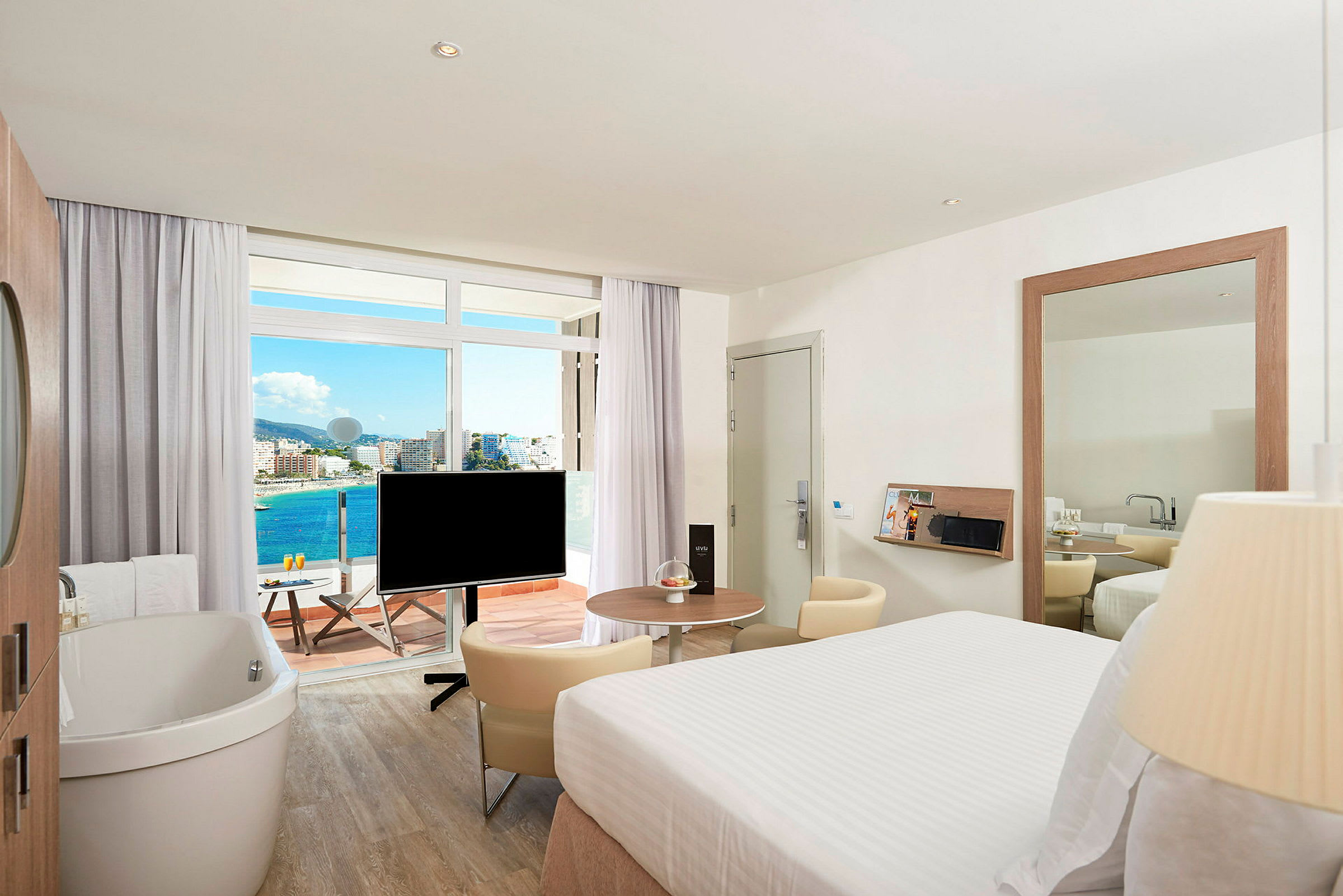 Ett hotellrum med en stor säng, en fristående badkar och en TV. Rummet har en balkong med utsikt över havet och staden. (AI-genererad text)