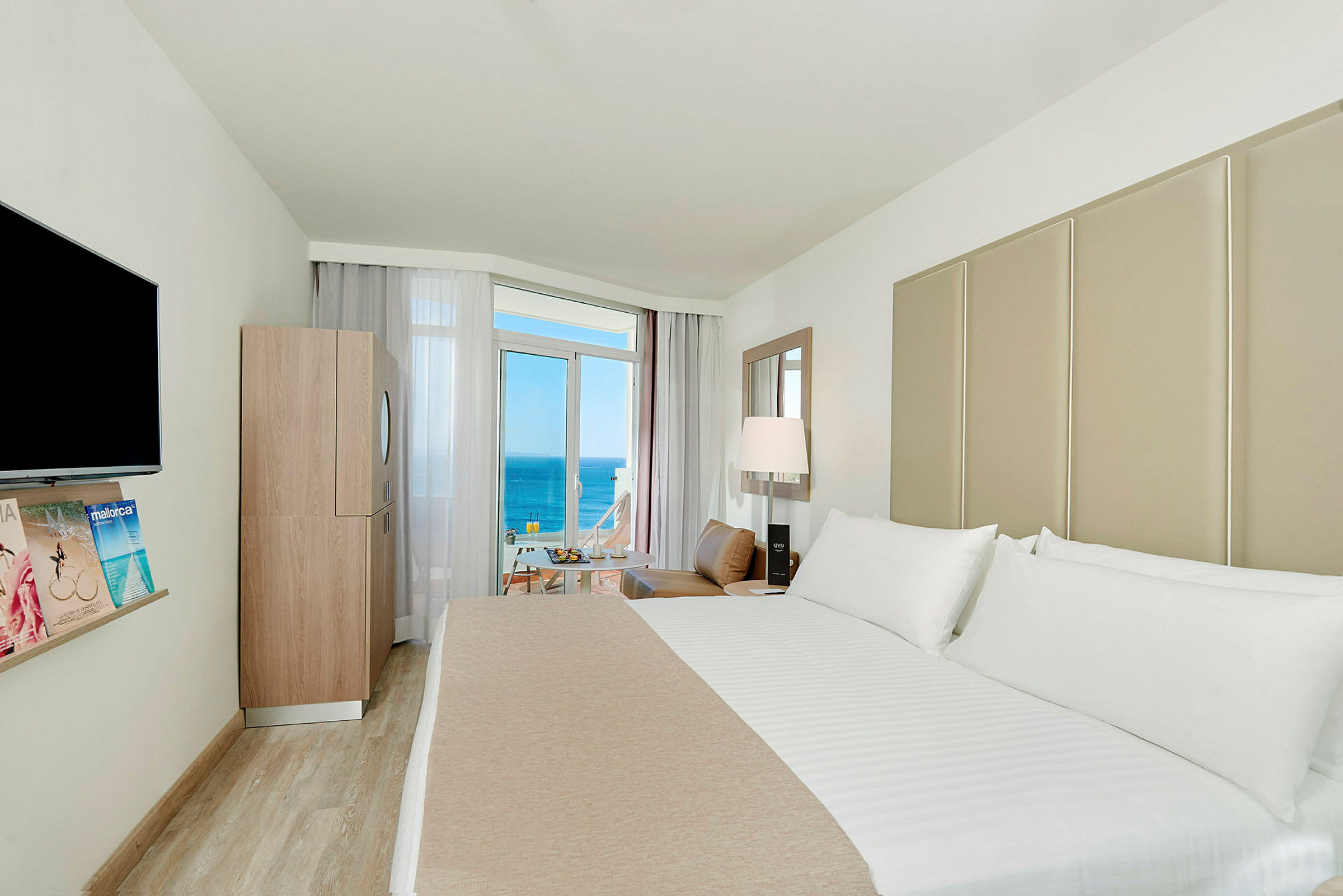 Ett hotellrum med en stor säng, en TV på väggen, en modern inredning och en balkong med utsikt över havet. (AI-genererad text)
