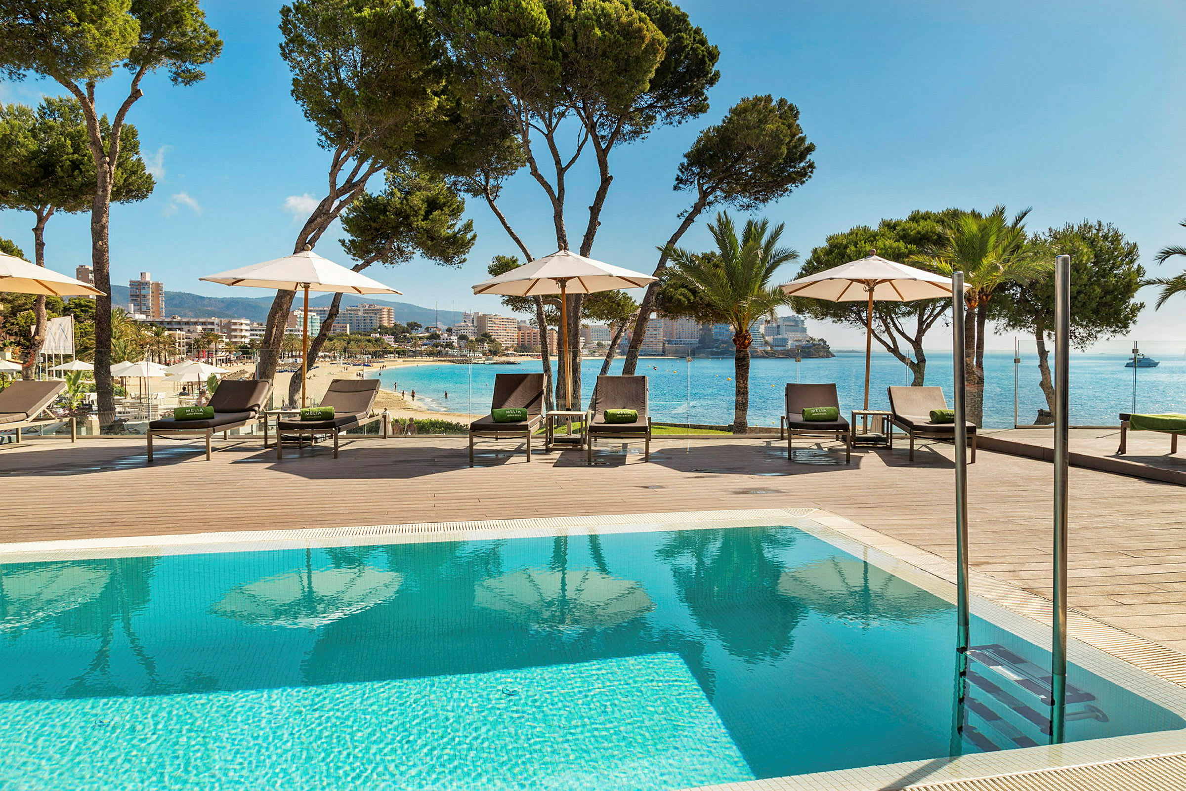 Bild av en pool med solstolar och parasoller vid en träterrass. I bakgrunden syns stranden, havet och en stad. (AI-genererad text)