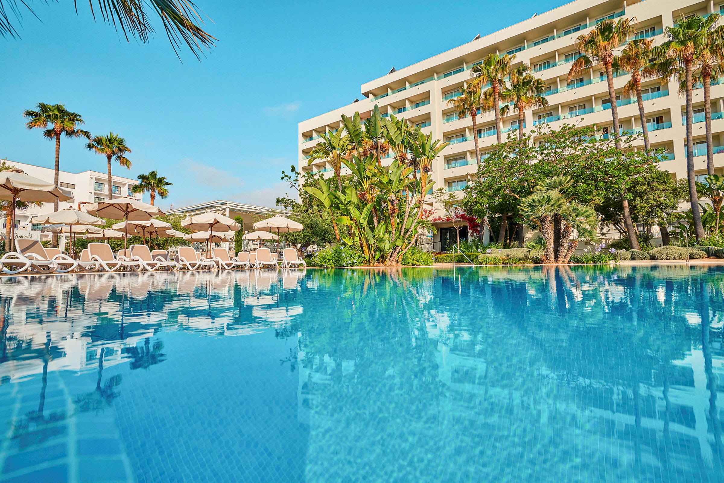 Hotell med pool och solstolar framför en trädgård och palmer. (AI-genererad text)