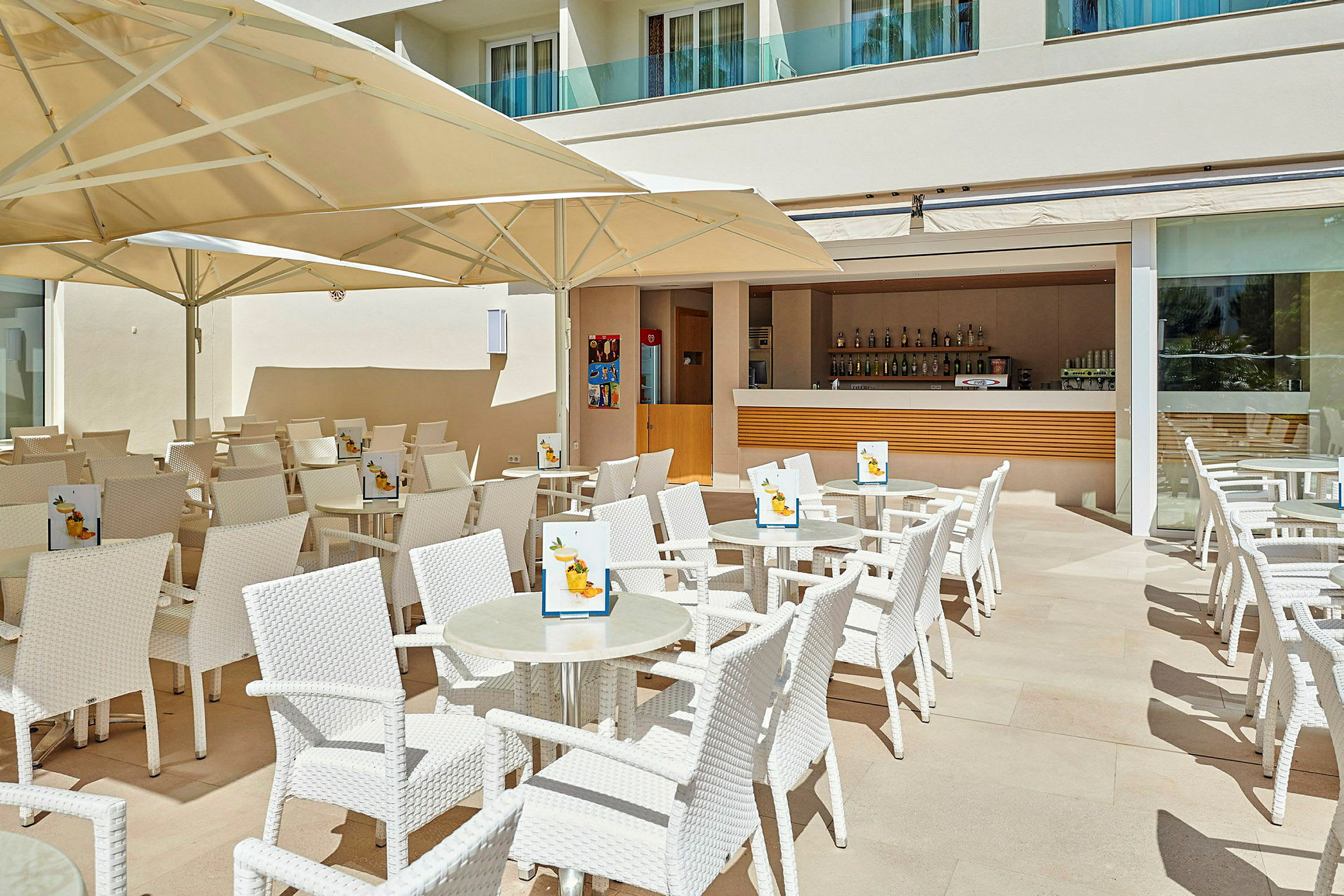 Terrass med vita stolar och bord, under stora parasoller, framför en öppen bar. (AI-genererad text)