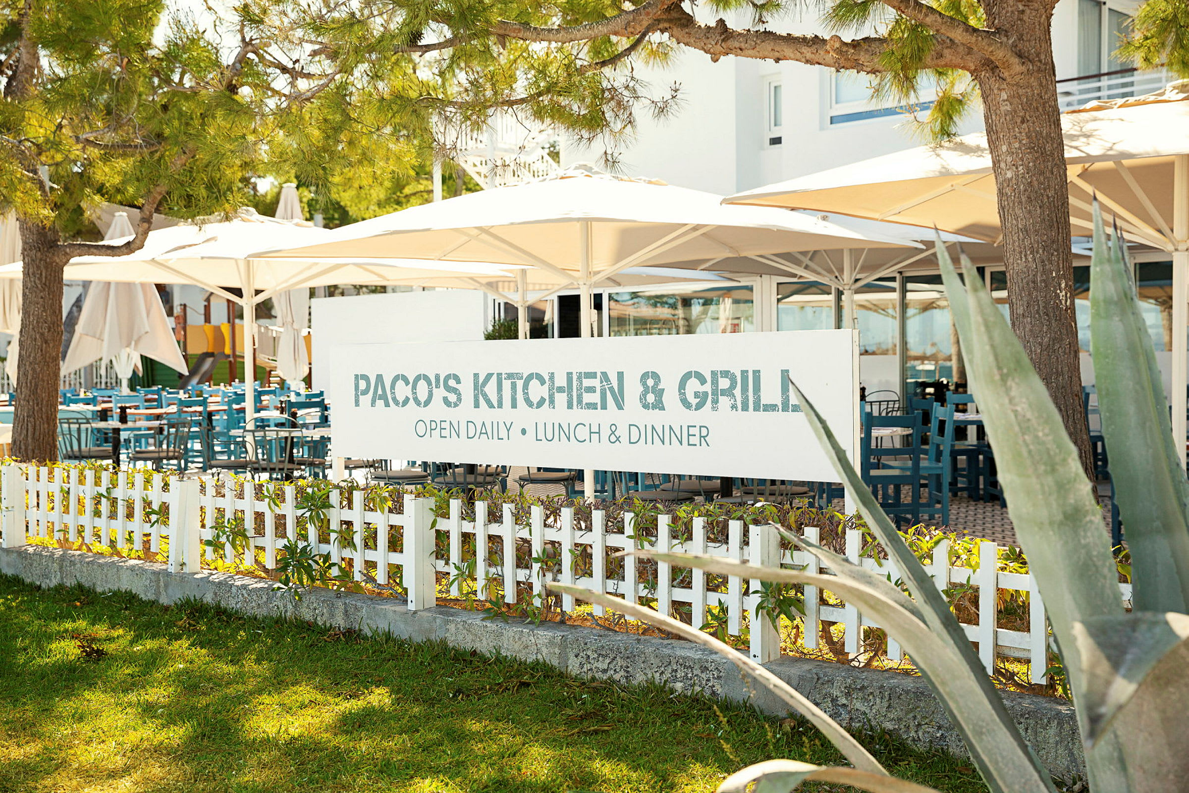 Skylt med texten 'Paco's Kitchen & Grill' framför en restaurang med uteservering. (AI-genererad text)