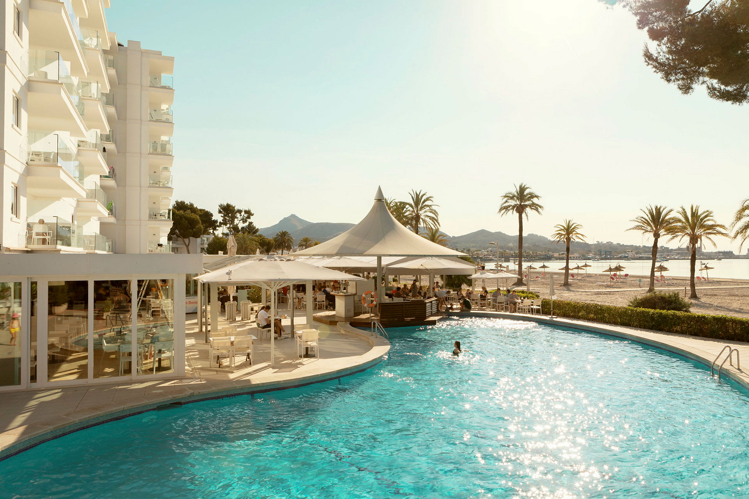 En bild av ett hotell med en pool i förgrunden och en sandstrand med palmer i bakgrunden. (AI-genererad text)