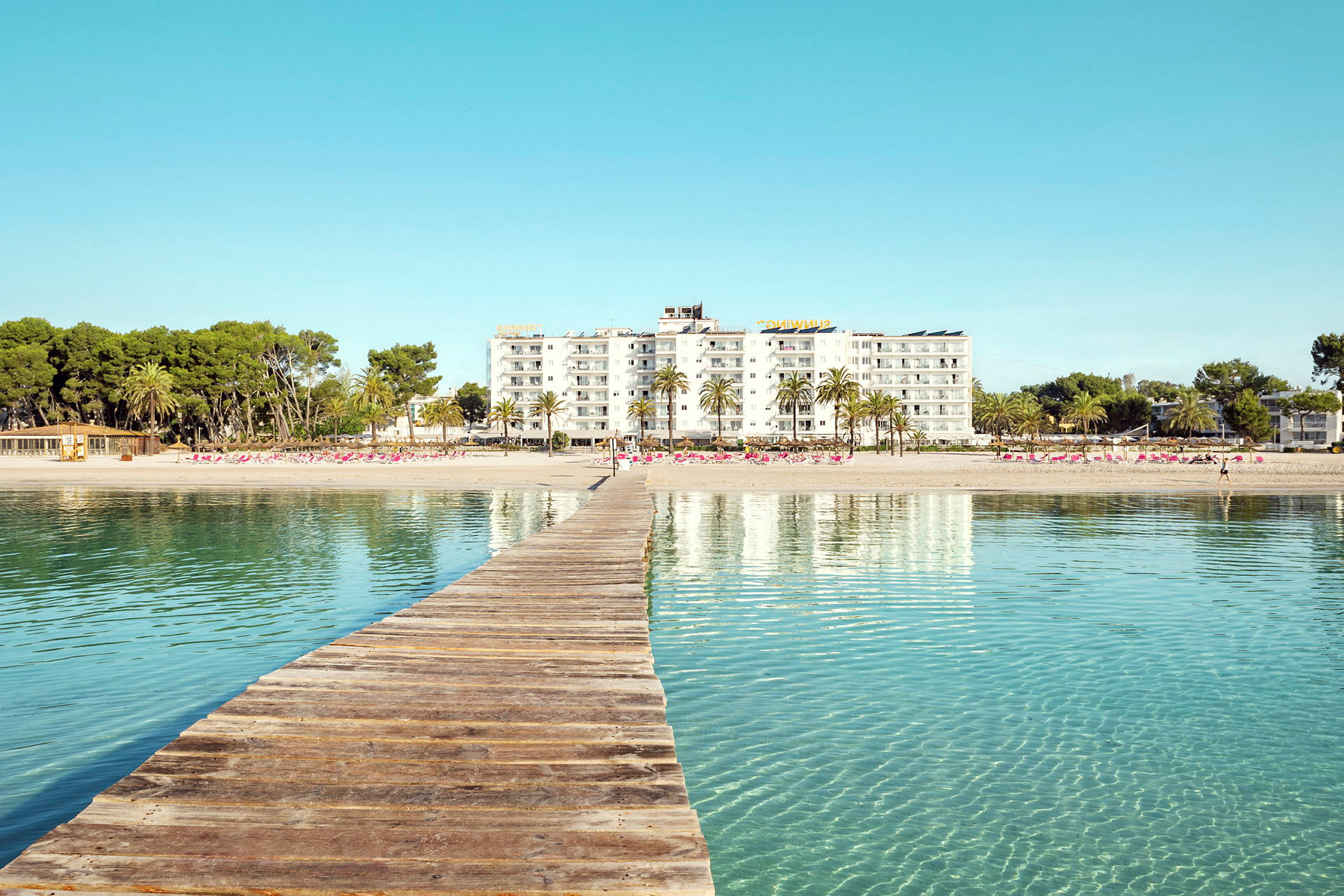 Träbrygga som leder mot en sandstrand med hotellbyggnader och palmer i bakgrunden. (AI-genererad text)