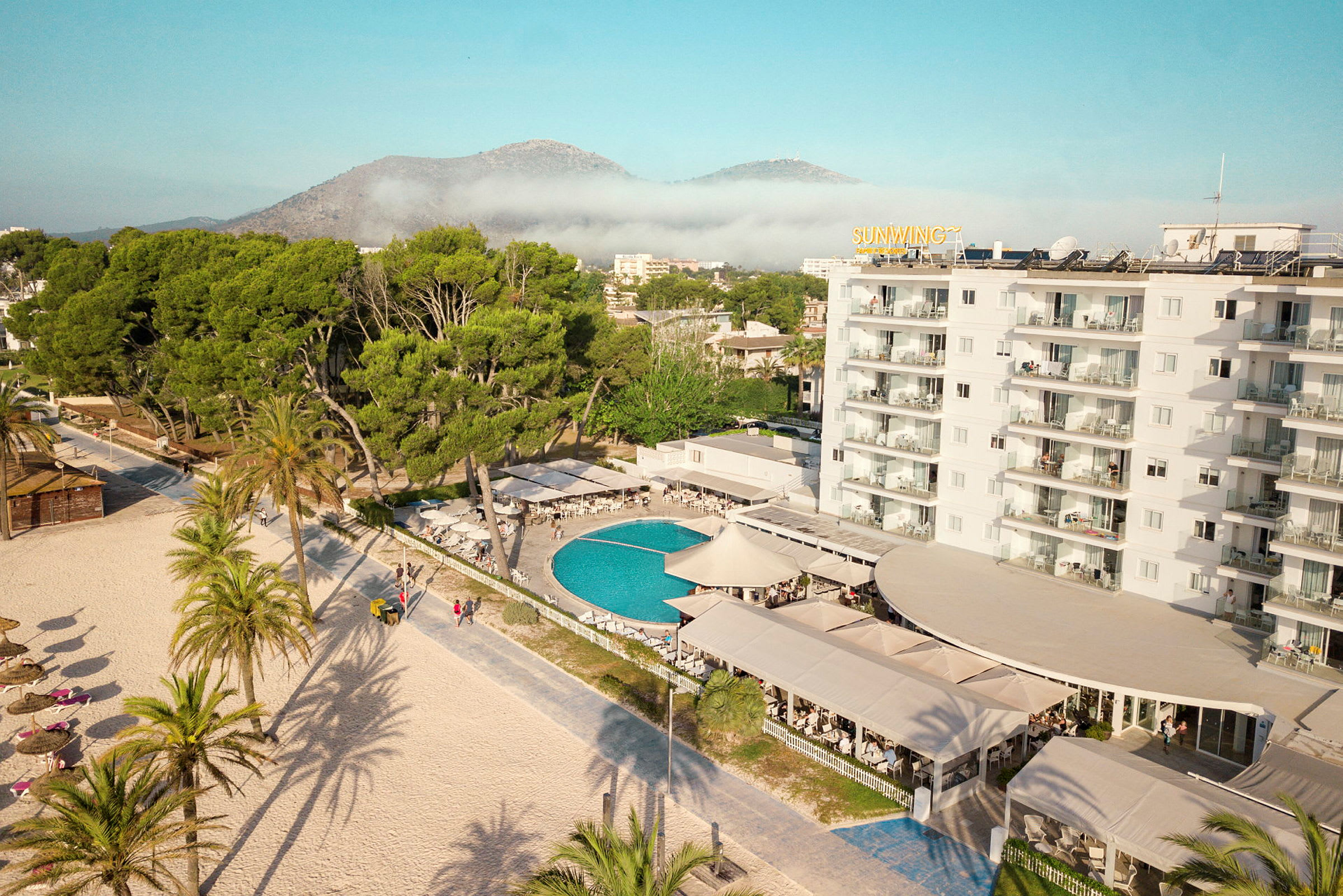 Hotelldel med pool, omgiven av grönska och med stranden i förgrunden. (AI-genererad text)