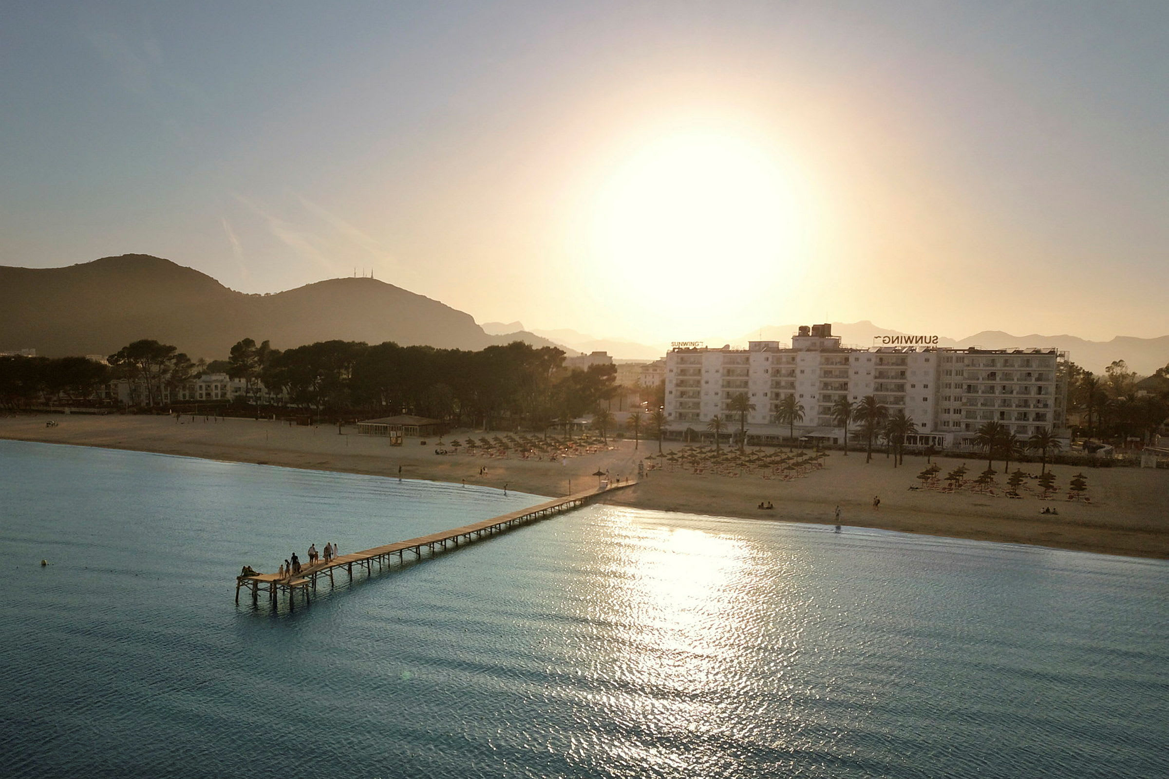 Strand med brygga, hotell och berg i bakgrunden vid solnedgång. (AI-genererad text)