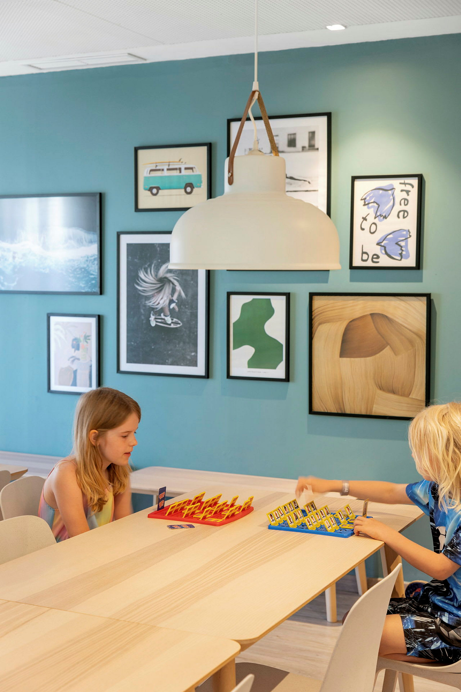 Två barn sitter vid ett bord och spelar ett brädspel, mot en turkos vägg med konst på. (AI-genererad text)