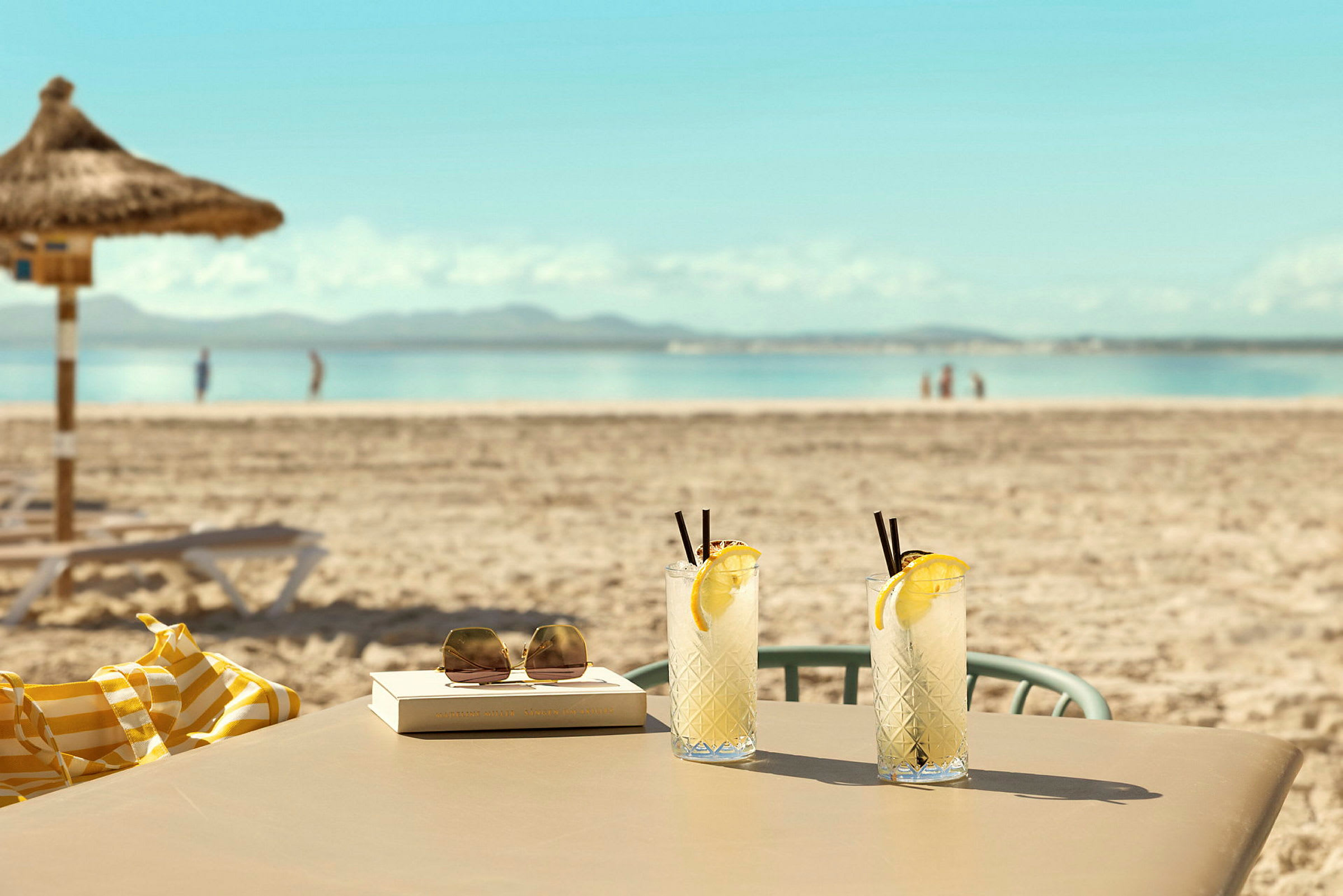 Två glas med lemonad står på ett bord vid en sandstrand med hav i bakgrunden. (AI-genererad text)
