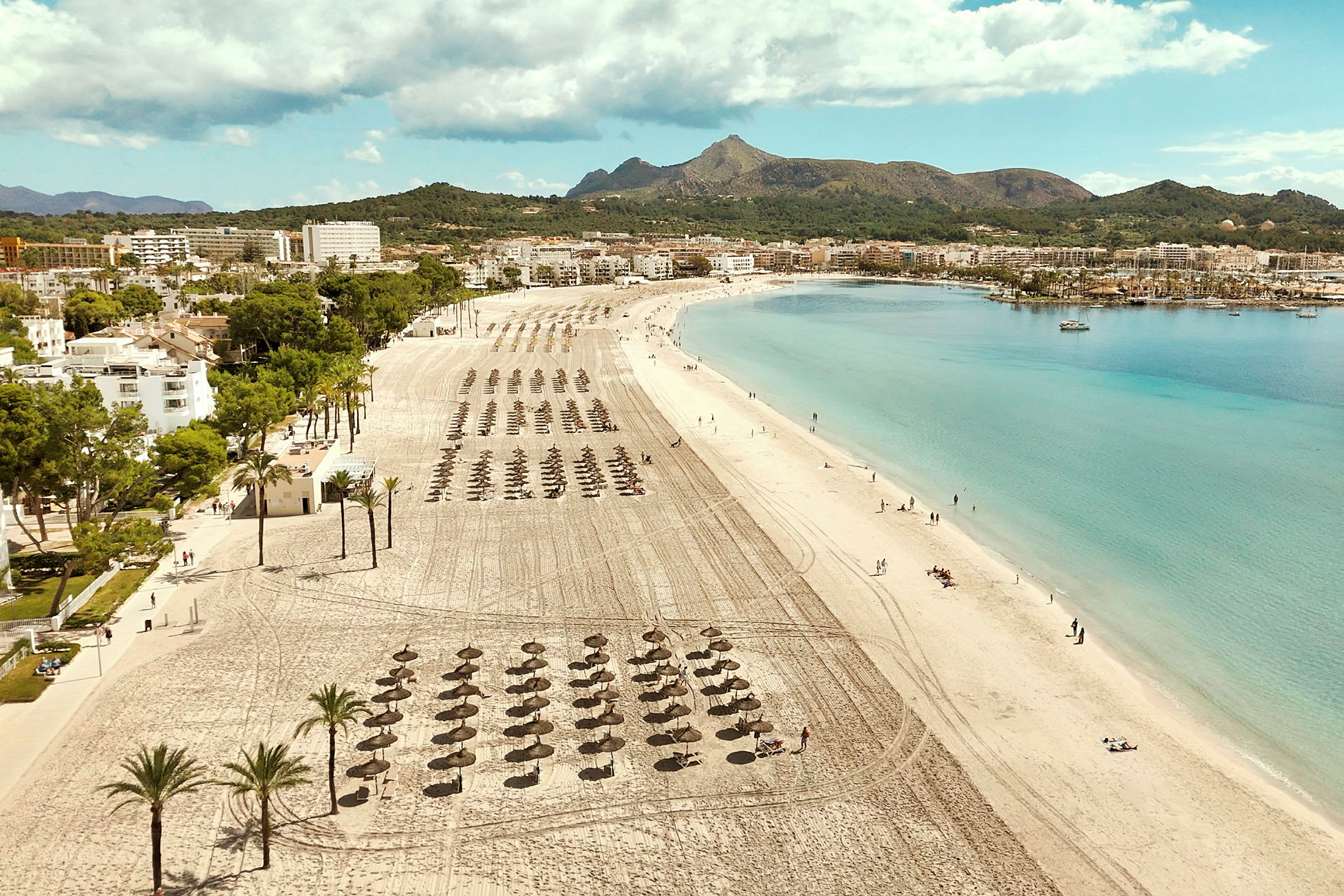 En långsträckt sandstrand med solparasoller och klarblått hav. I bakgrunden syns byggnader och grönskande berg. (AI-genererad text)