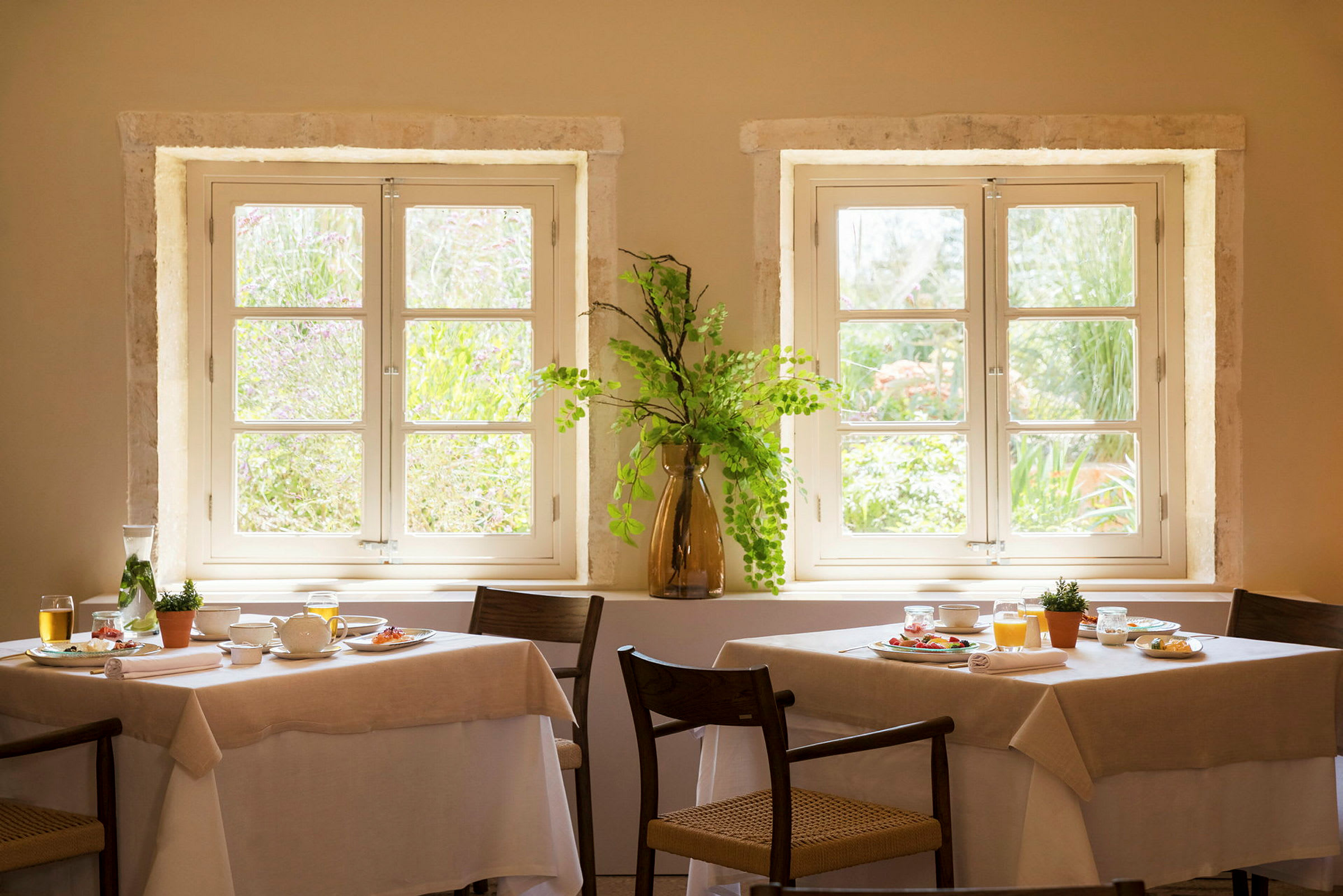 Två dukade bord framför soliga fönster med en vas med gröna kvistar. (AI-genererad text)