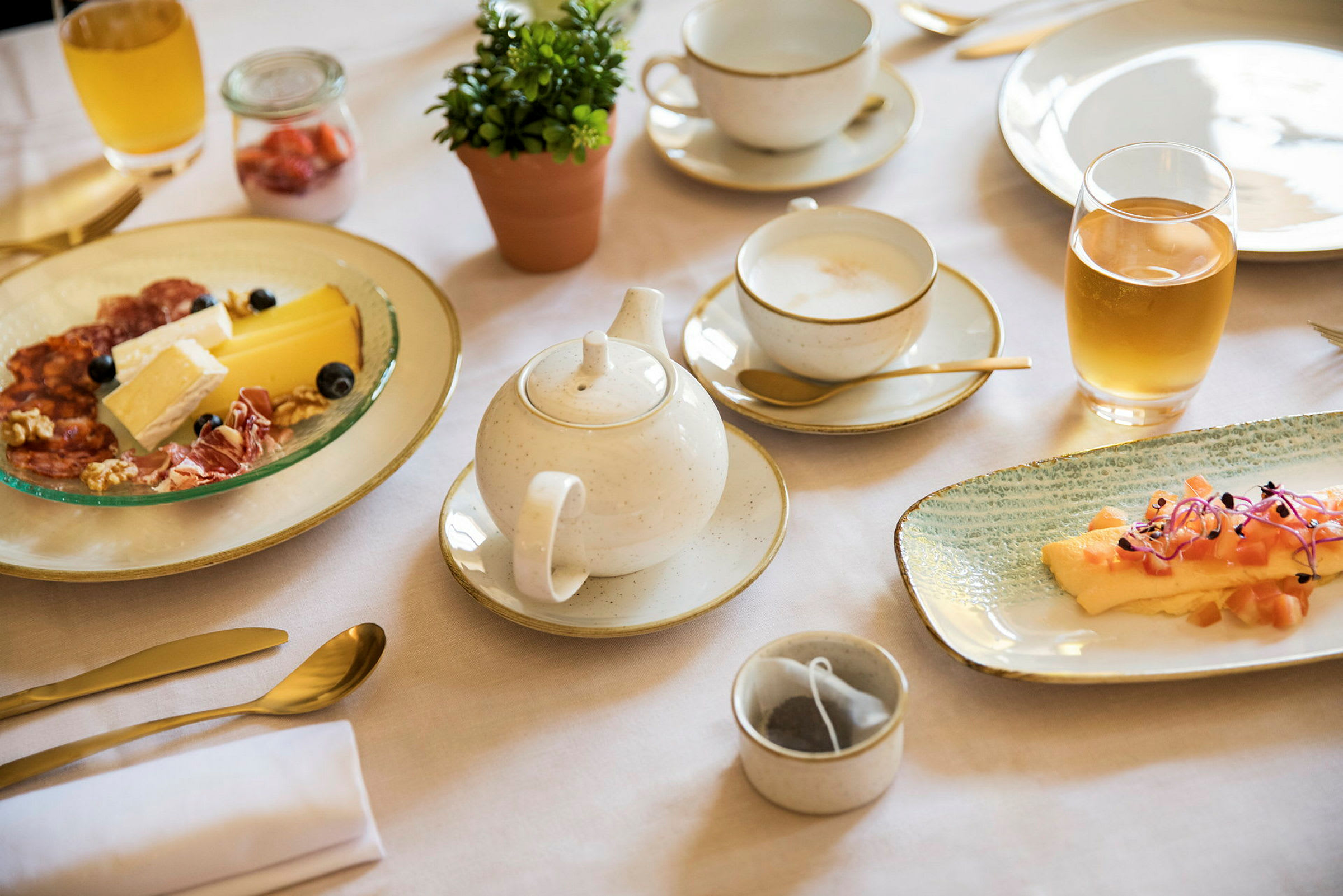 Frukost med olika rätter, inklusive ost och charkuterier, en liten växt i kruka och te serverat i en kopp. (AI-genererad text)