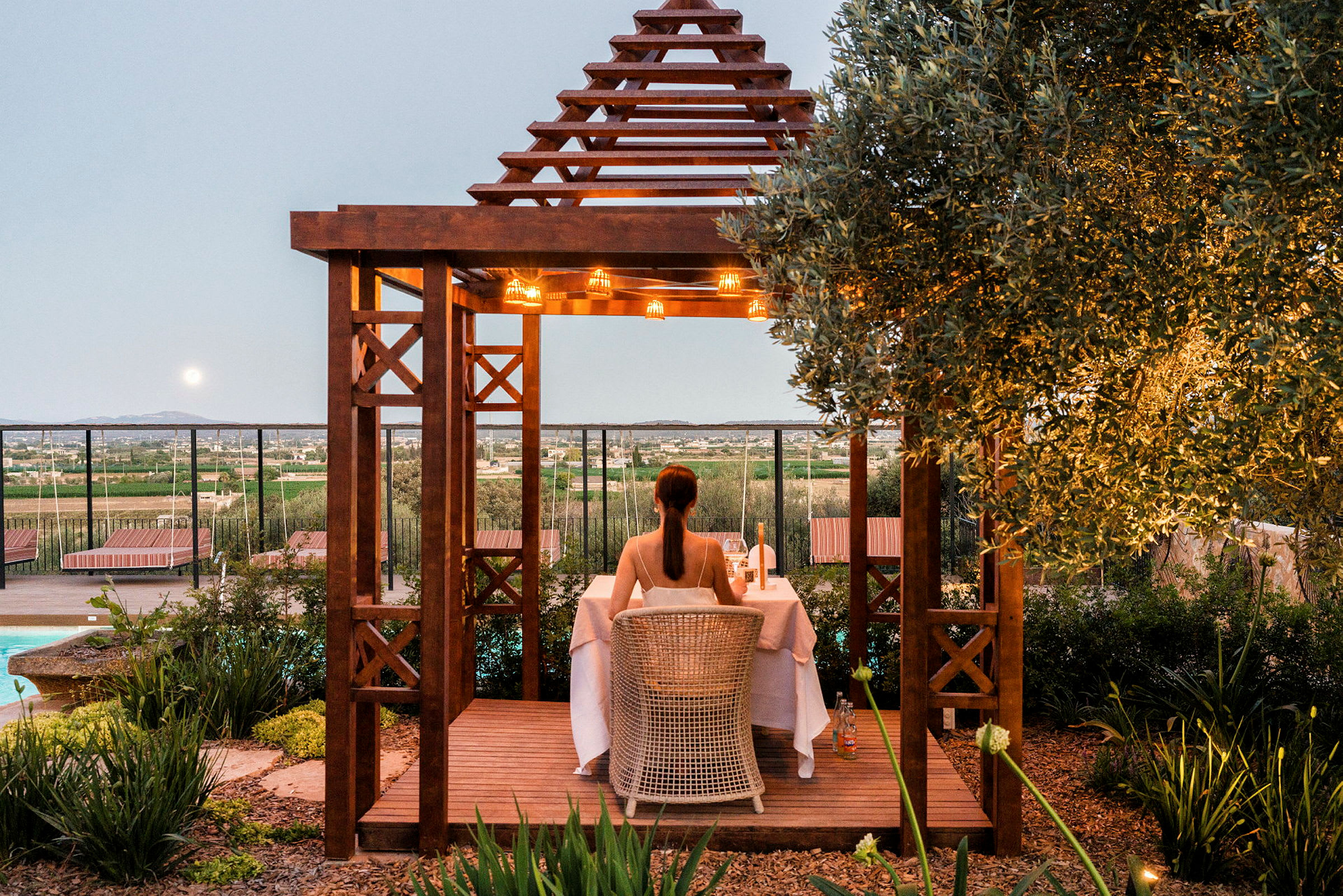En person sitter vid ett bord under en träpergola som är dekorerad med ljus, omgiven av grönska med utsikt över landskapet. (AI-genererad text)
