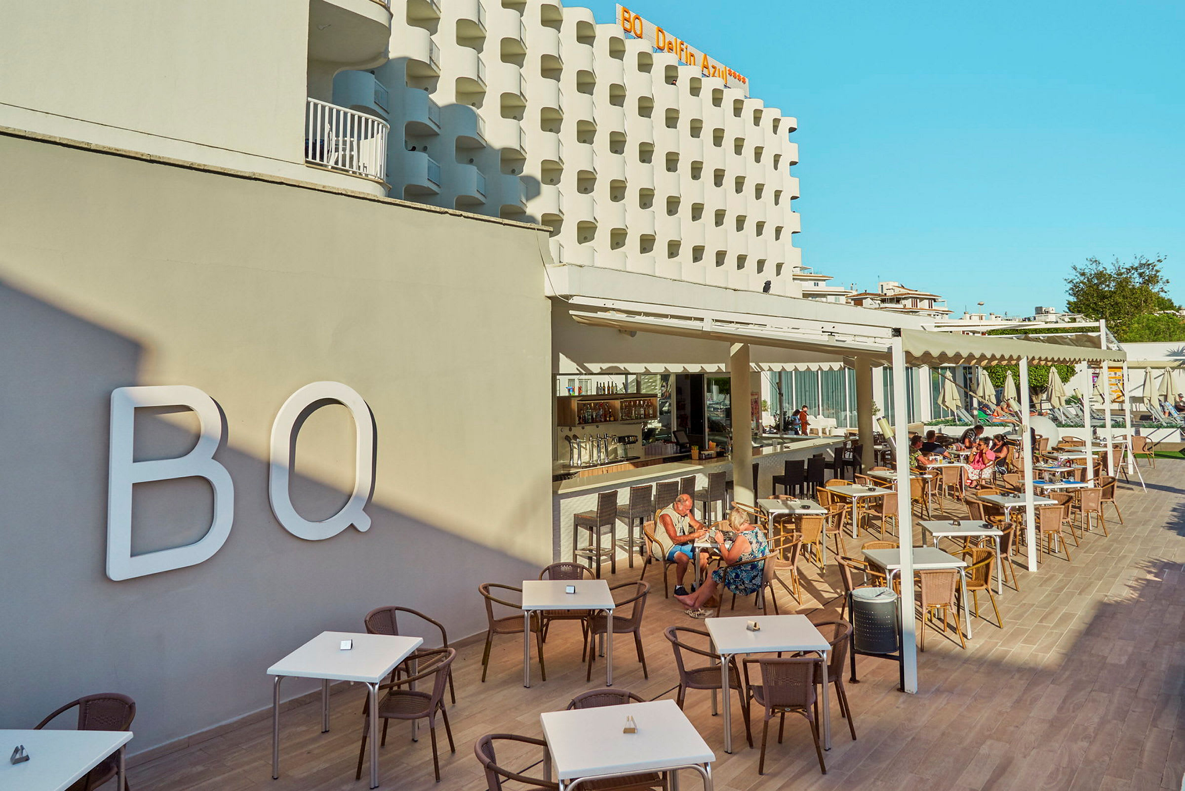 Hotellfasad och utomhusservering med bord och stolar, människor som sitter och äter. (AI-genererad text)