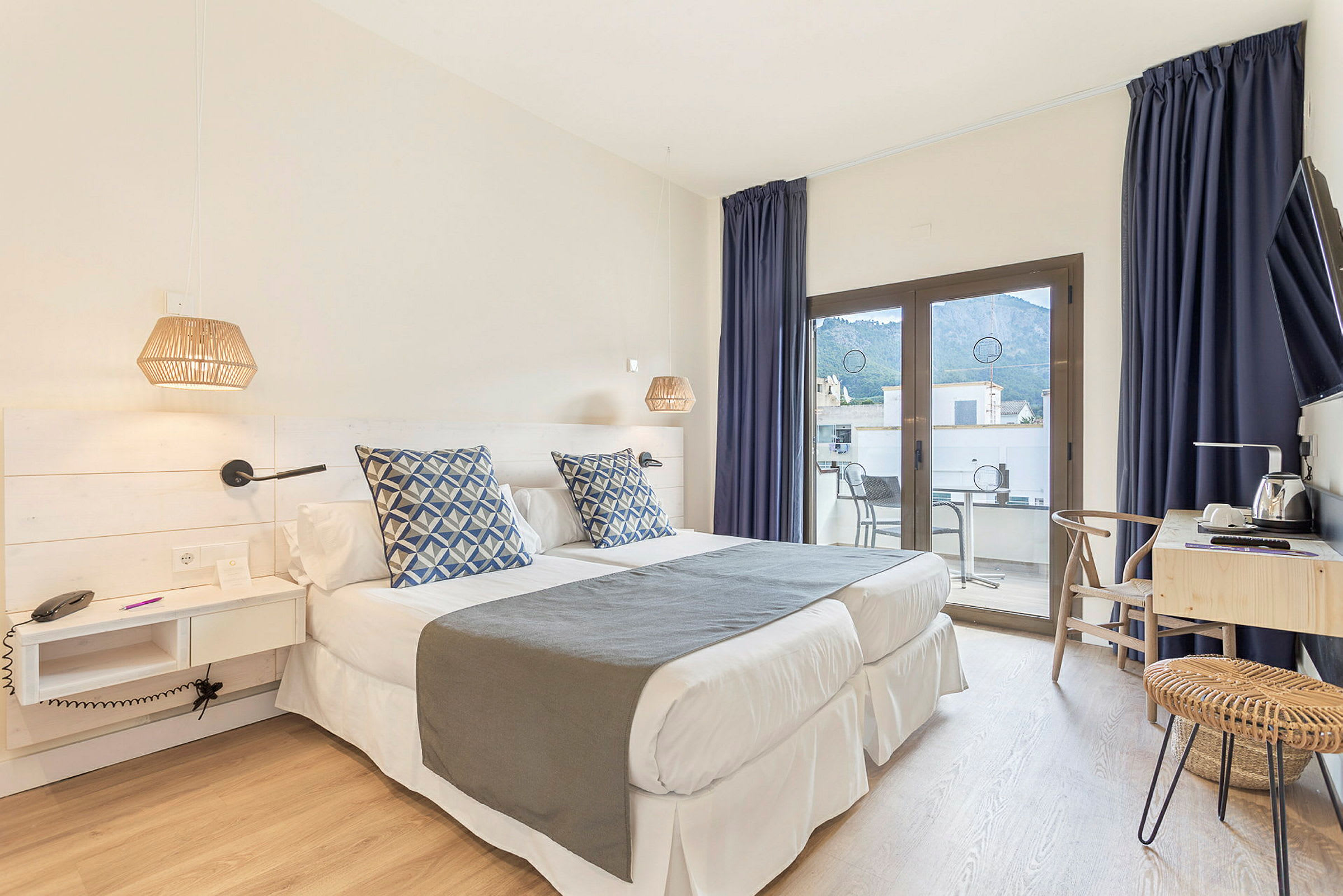 Ett hotellrum med två enkelsängar, blå dekorkuddar, skrivbord och en balkong med utsikt. (AI-genererad text)