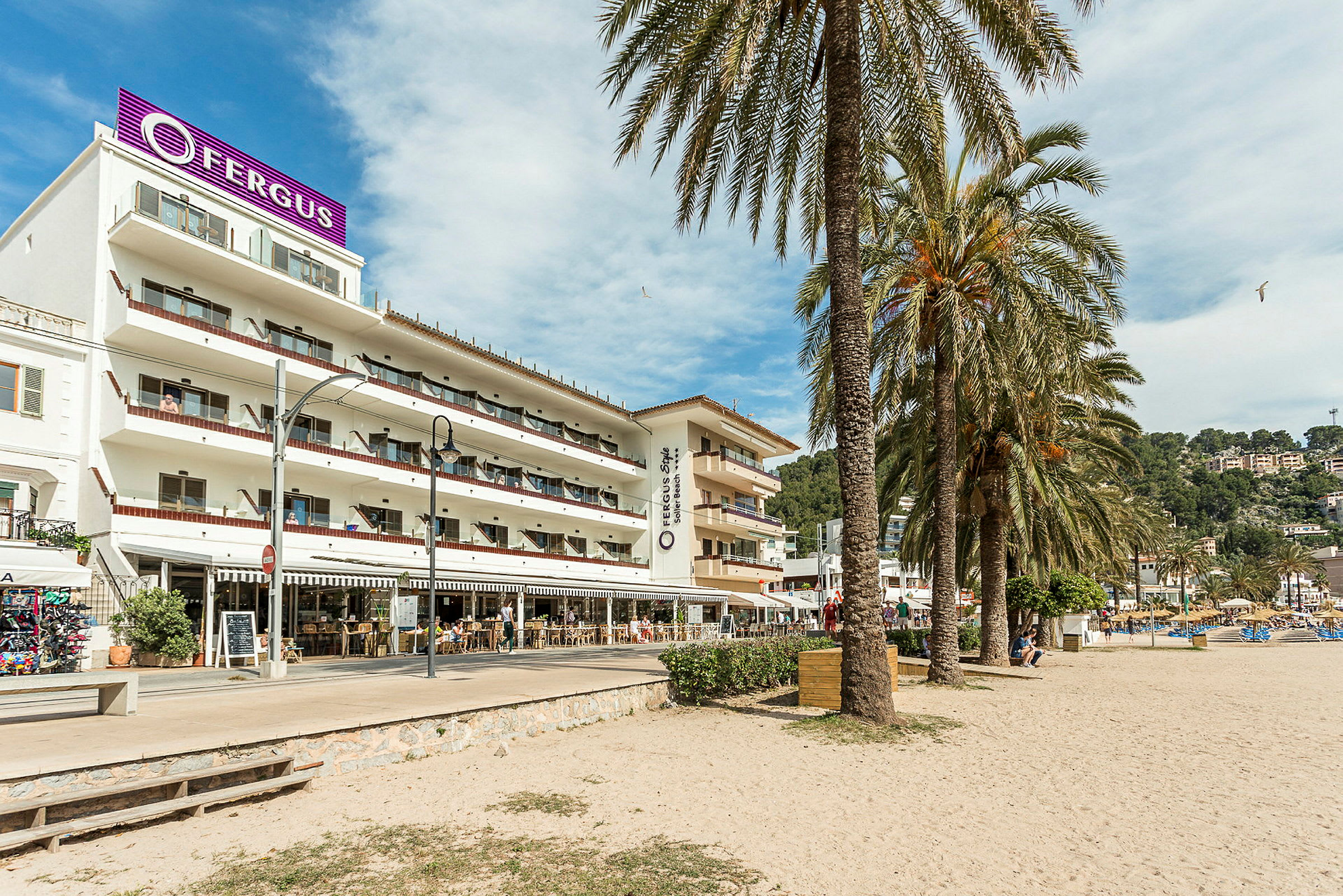 Ett hotell med flera våningar och balkonger ligger vid en sandstrand, omgiven av palmer. (AI-genererad text)