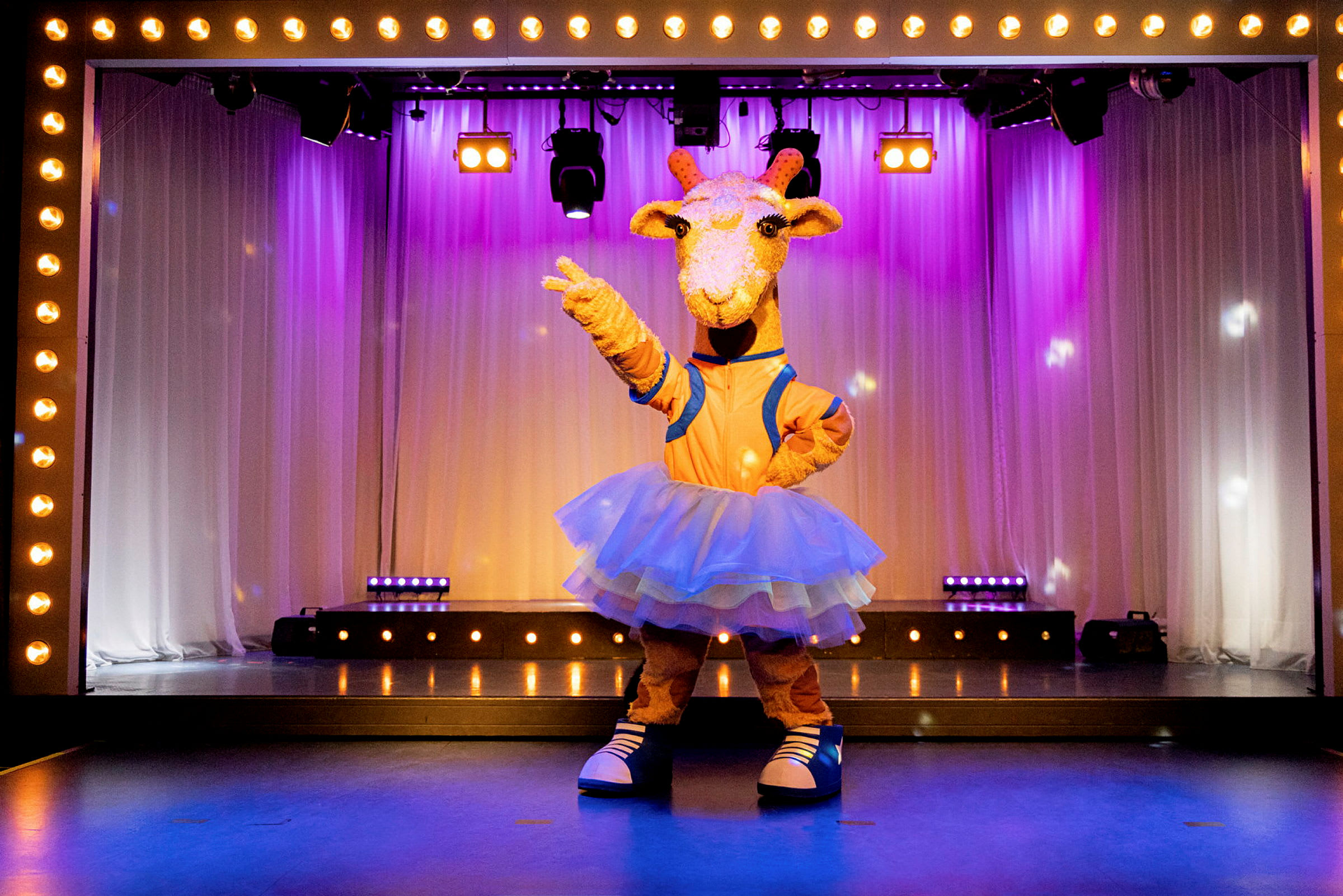 En giraffklädd figur står på en scen dekorerad med ljus och vita draperier. (AI-genererad text)