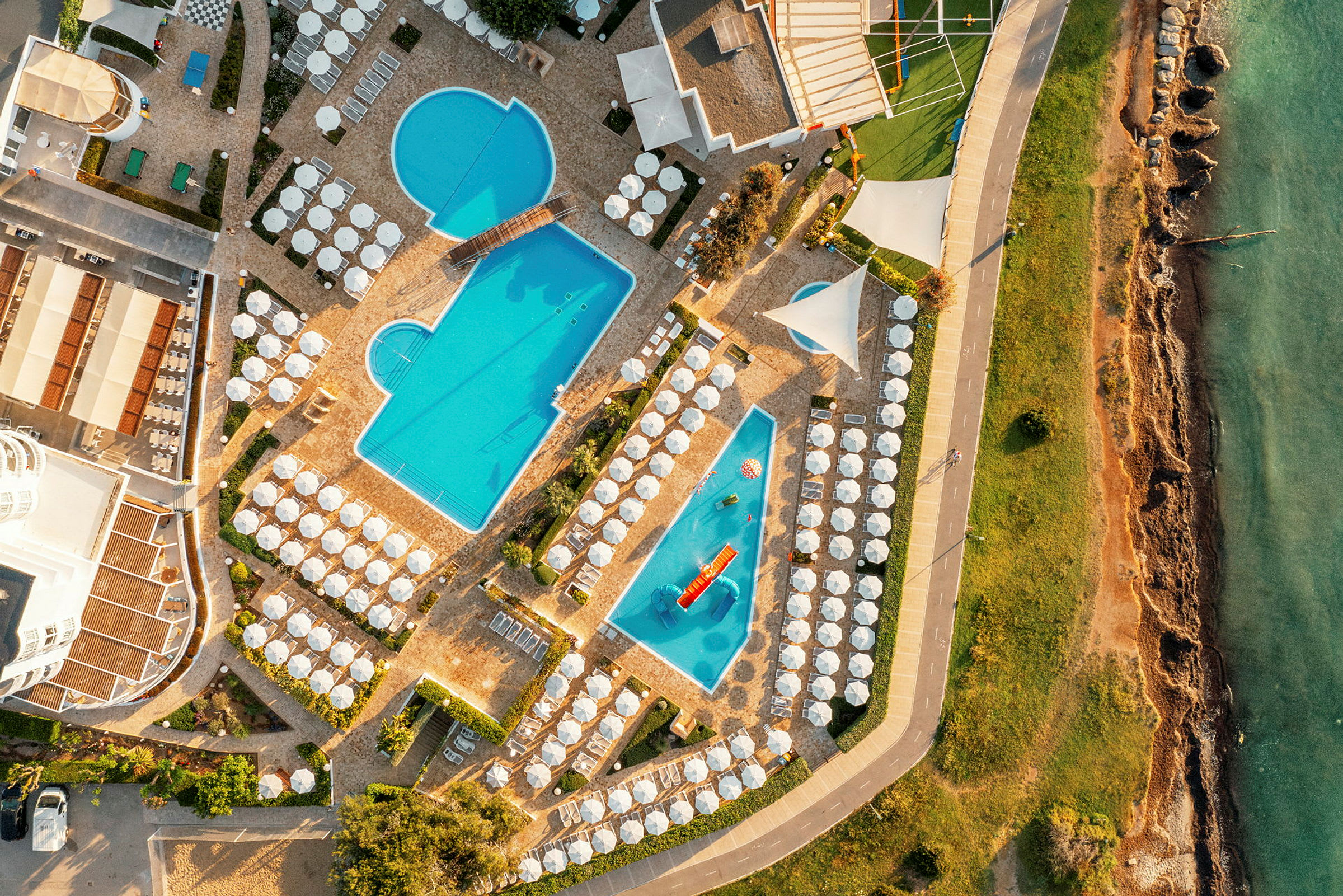 Flygbild av hotellpoolområde med solstolar och parasoller nära havet. (AI-genererad text)