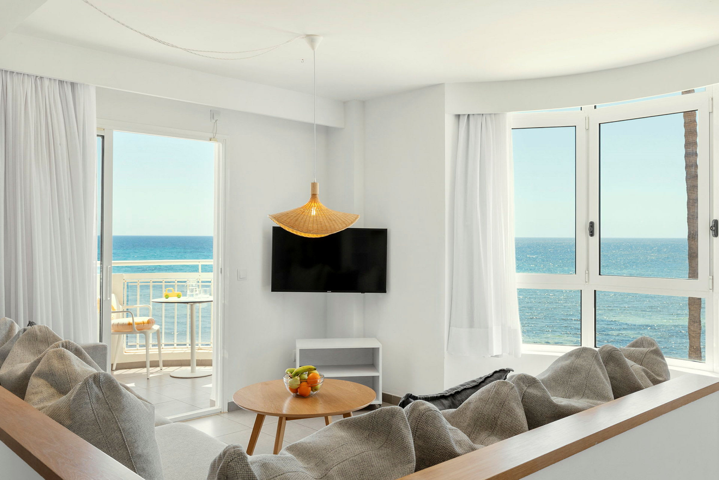 Ett ljust vardagsrum med soffor, ett bord med fruktskål, en TV, stora fönster och en balkong med utsikt över havet. (AI-genererad text)
