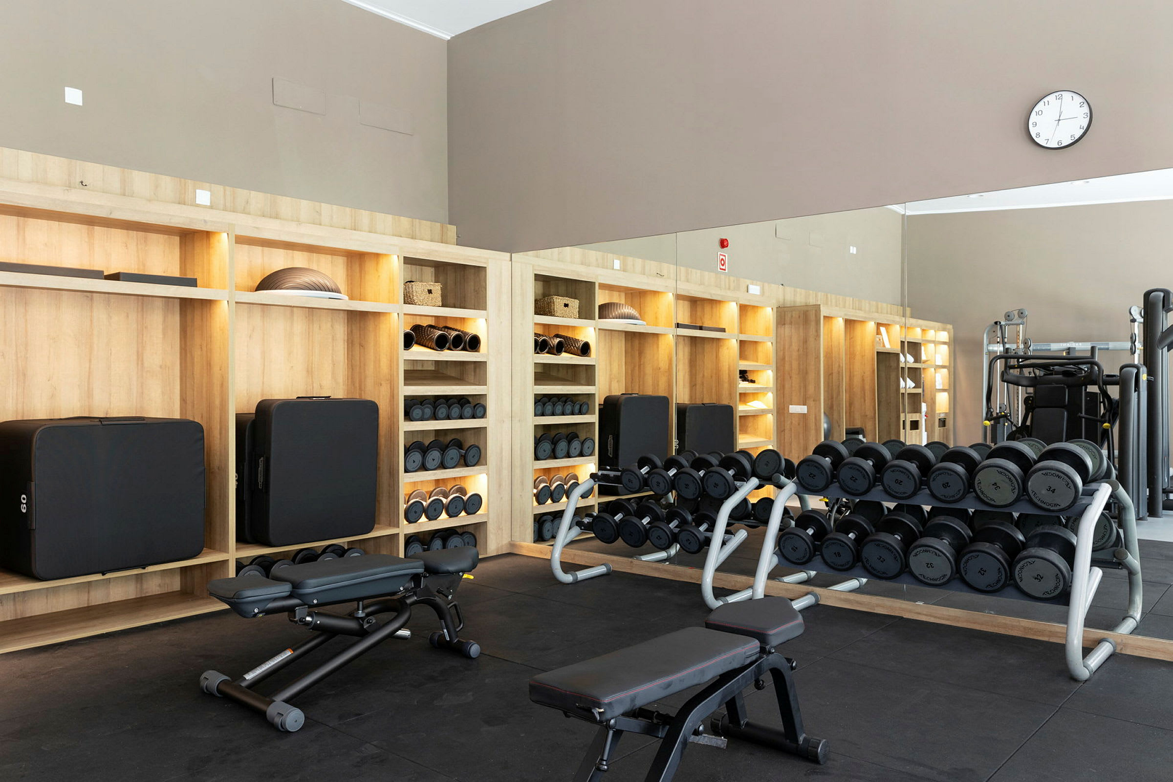 Ett gym med hyllor fyllda med träningsutrustning och vikter. Bänkar och speglar finns på platset. (AI-genererad text)