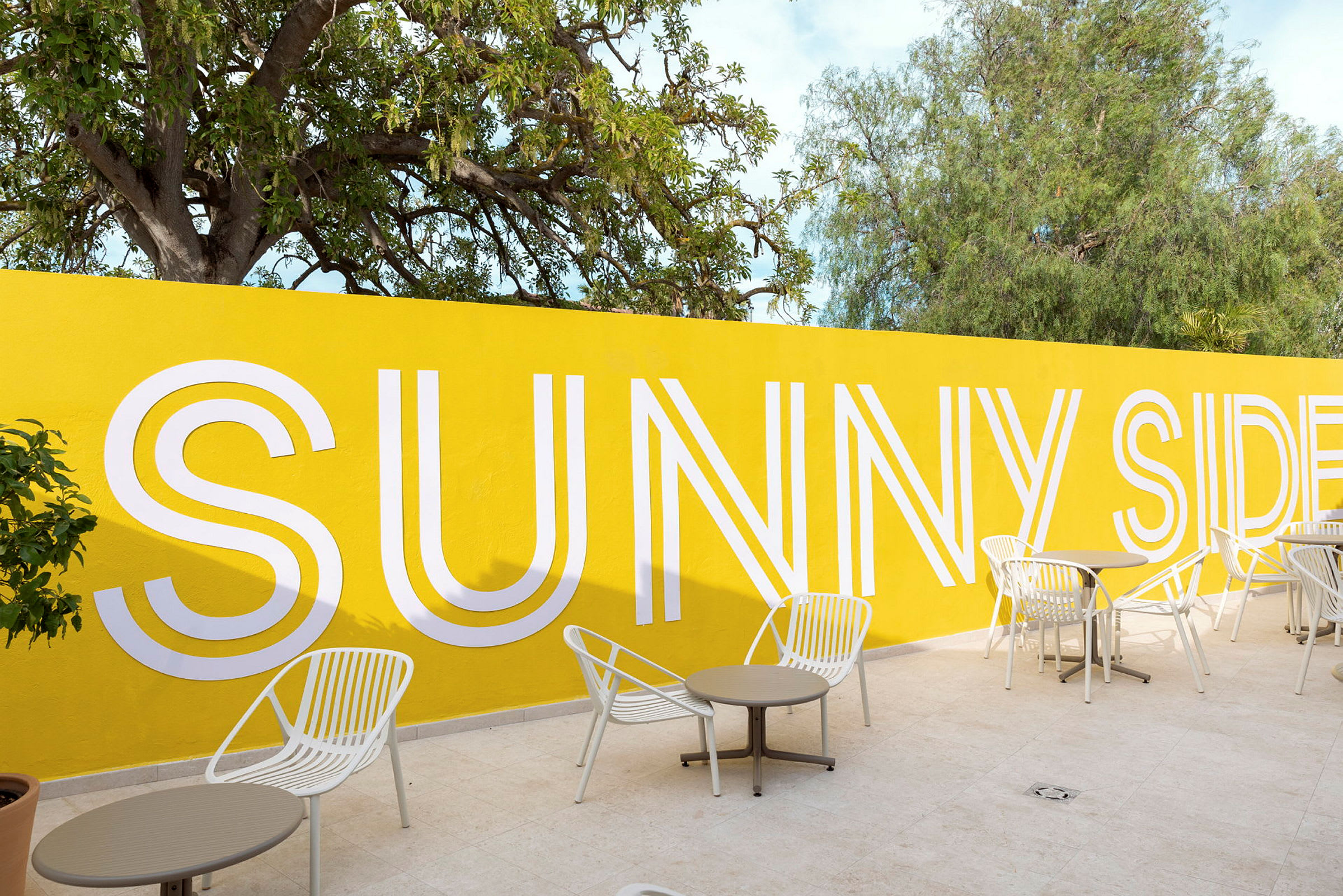 En gul vägg med texten 'Sunny Side' i vitt, framför står vita stolar och bord på en ljus stenbelagd yta. (AI-genererad text)
